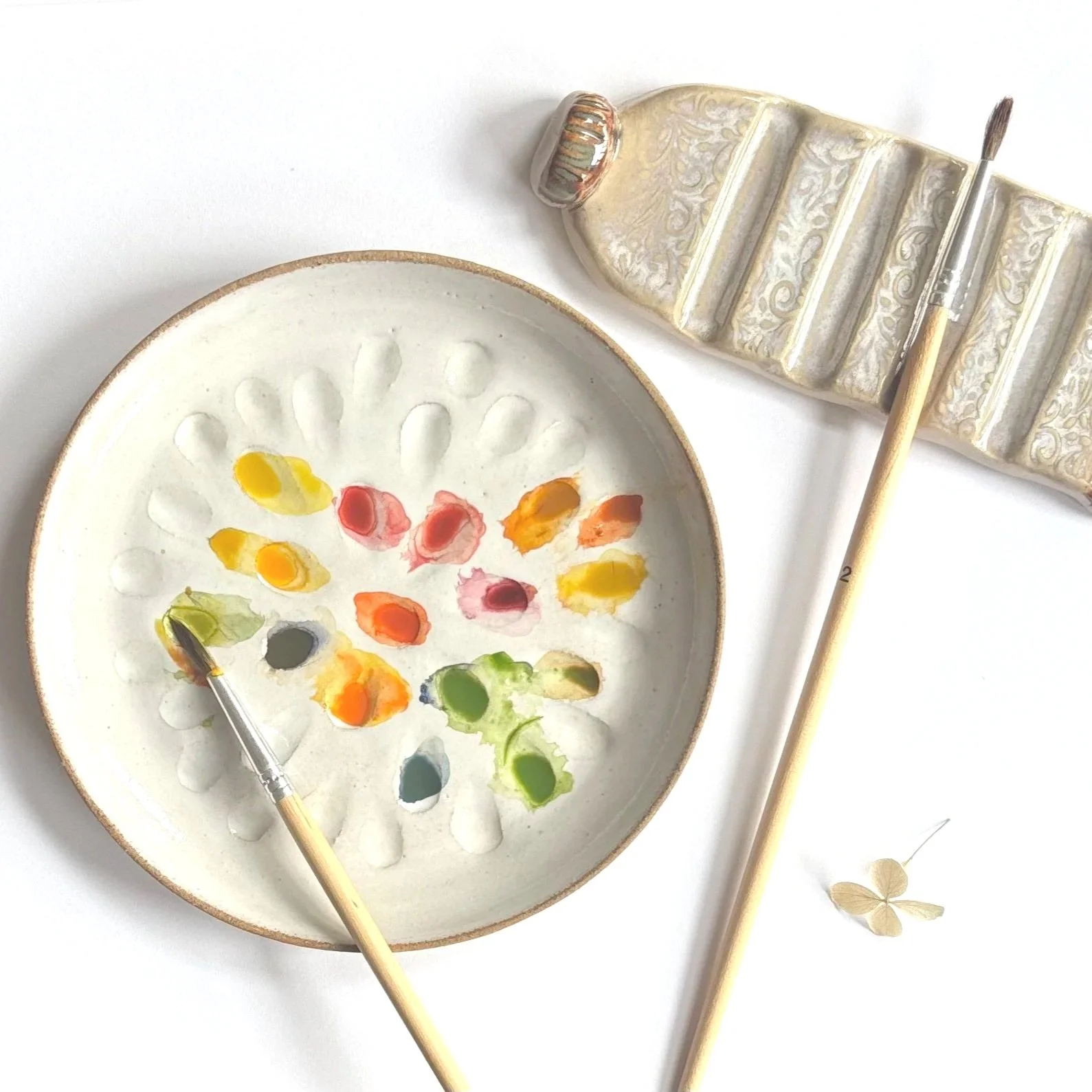 Three paintbrushes resting on a decorative ceramic brush holder with a lid, and a small yellow flower nearby.