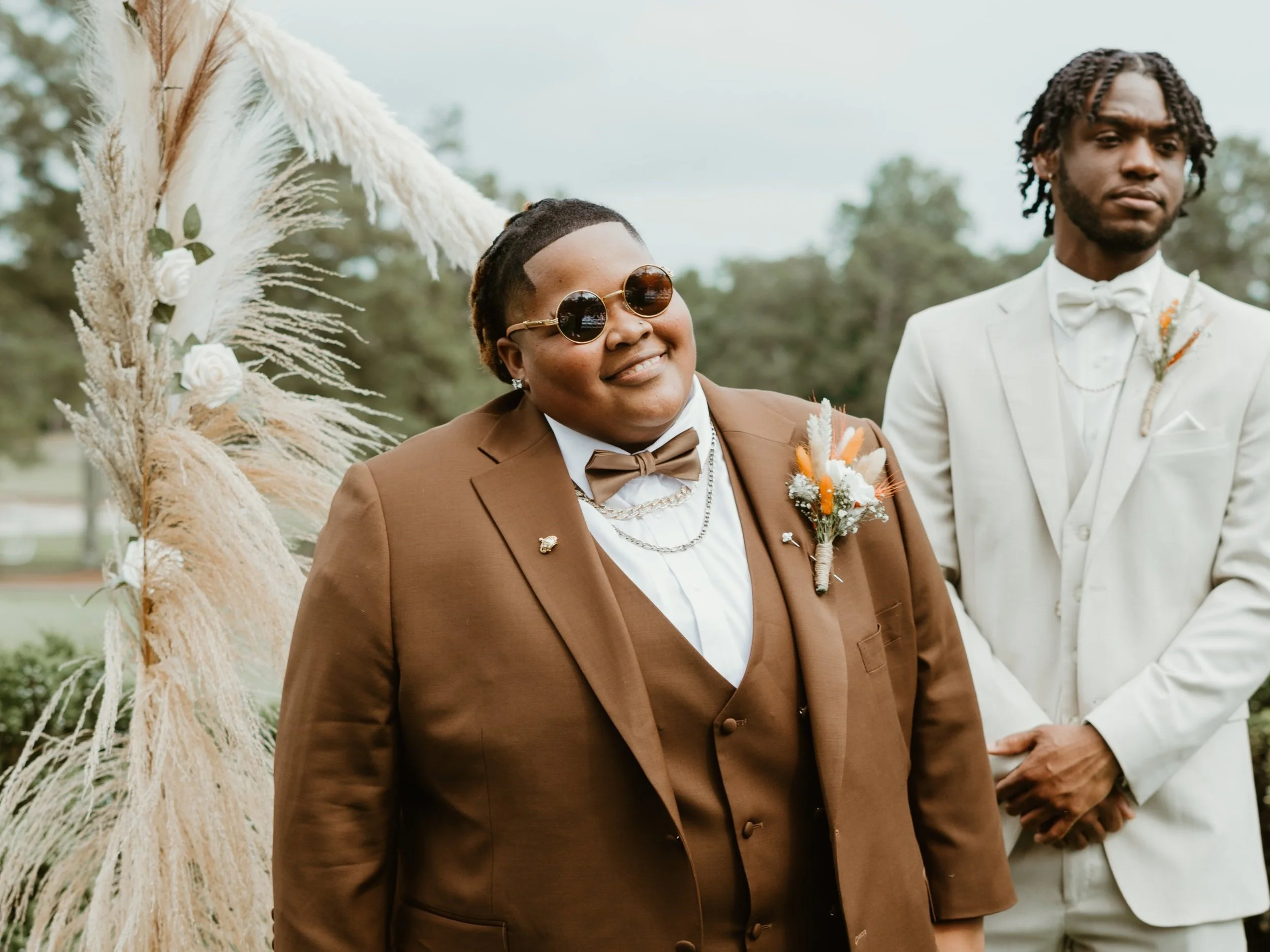 Two people in formal attire; one wearing a brown suit and sunglasses, and the other in a cream suit, both with boutonnières, standing outdoors.