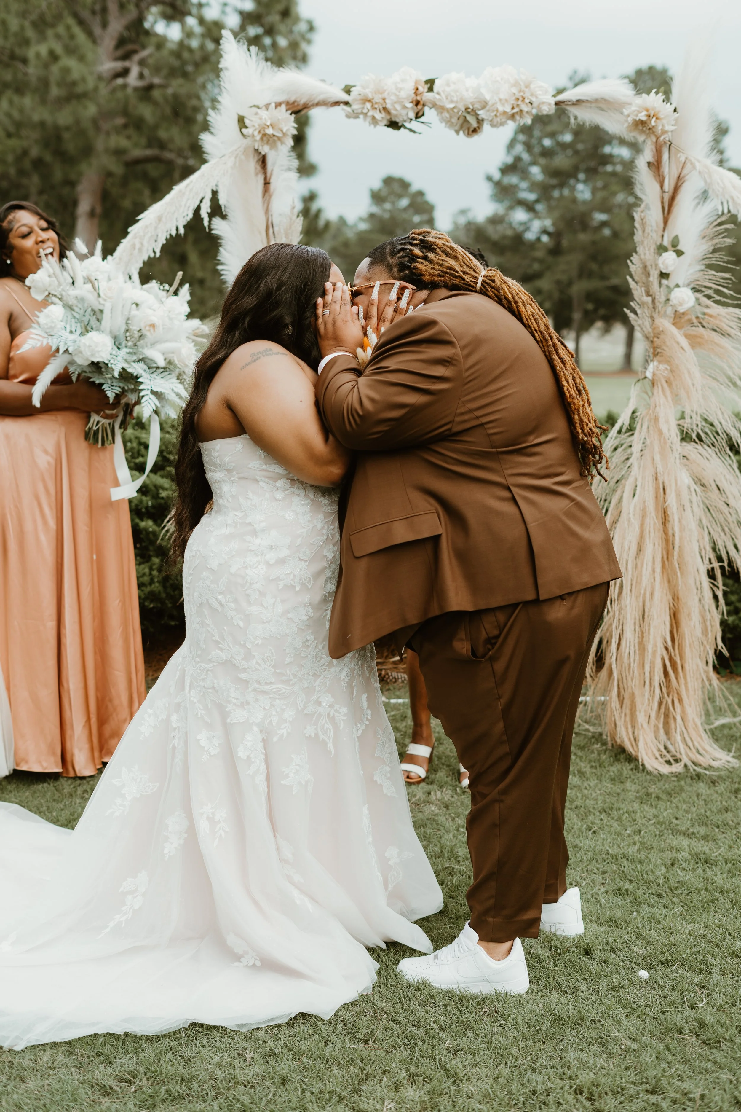 A couple getting married outdoors under a floral arch, wearing a white gown and brown suit. A bridesmaid in a peach dress stands nearby holding a bouquet. The setting is lush with greenery.