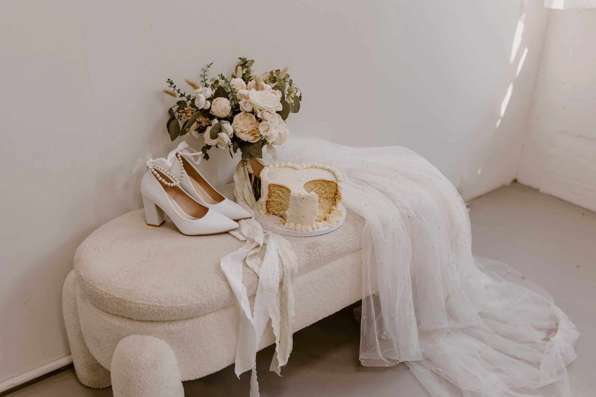 Wedding setup with white high-heeled shoes, floral bouquet, sliced cake, and tulle fabric on a bench.