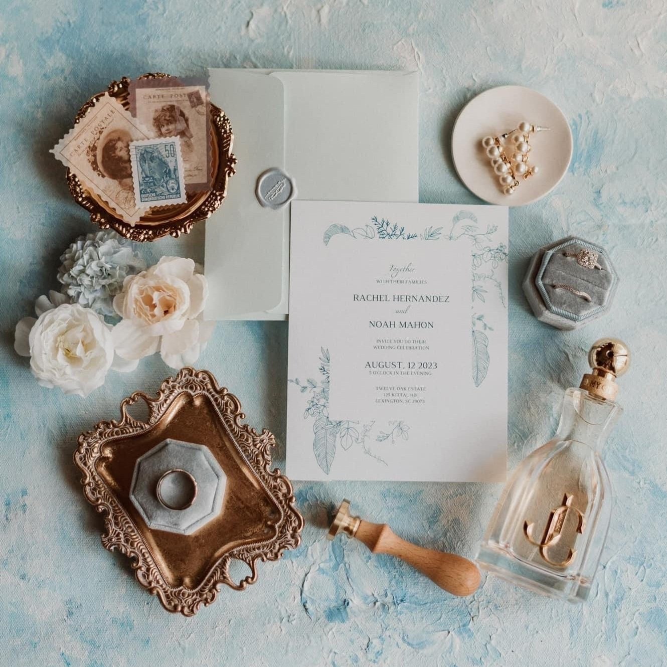 Flat lay of wedding invitation suite with vintage stamps, flowers, pearl earrings, blue jewelry box, gold tray, and perfume bottle on textured background.