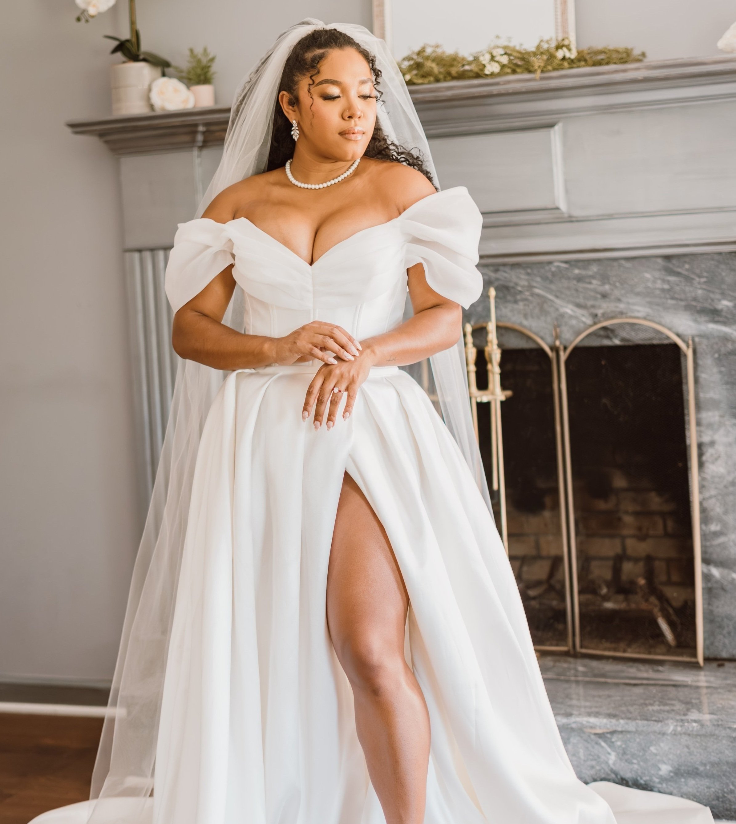 Bride in elegant white wedding dress with off-shoulder sleeves and a slit, standing in front of a fireplace with flowers.