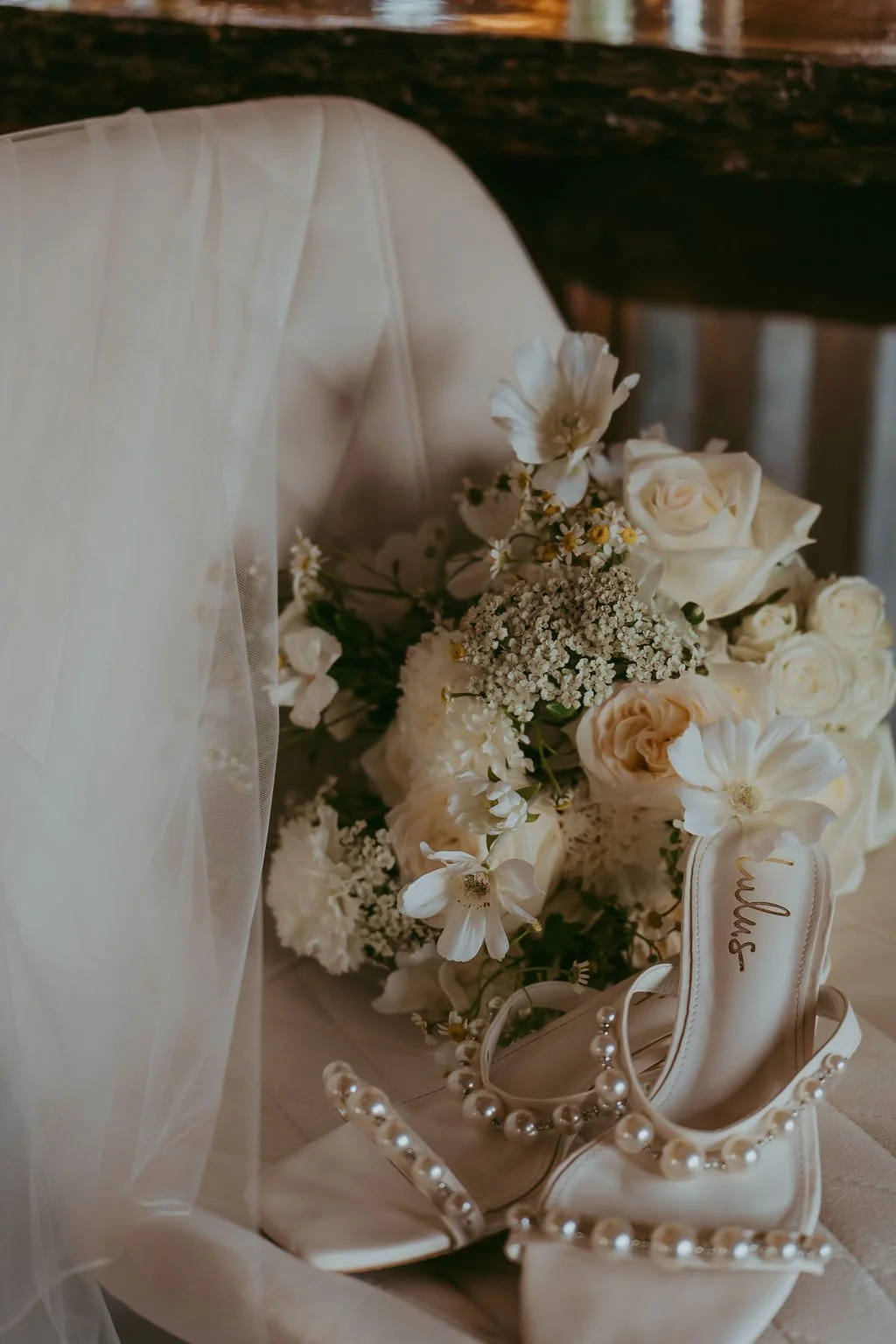 Bridal bouquet with white flowers, pearl-embellished shoes, and a veil draped over a chair.