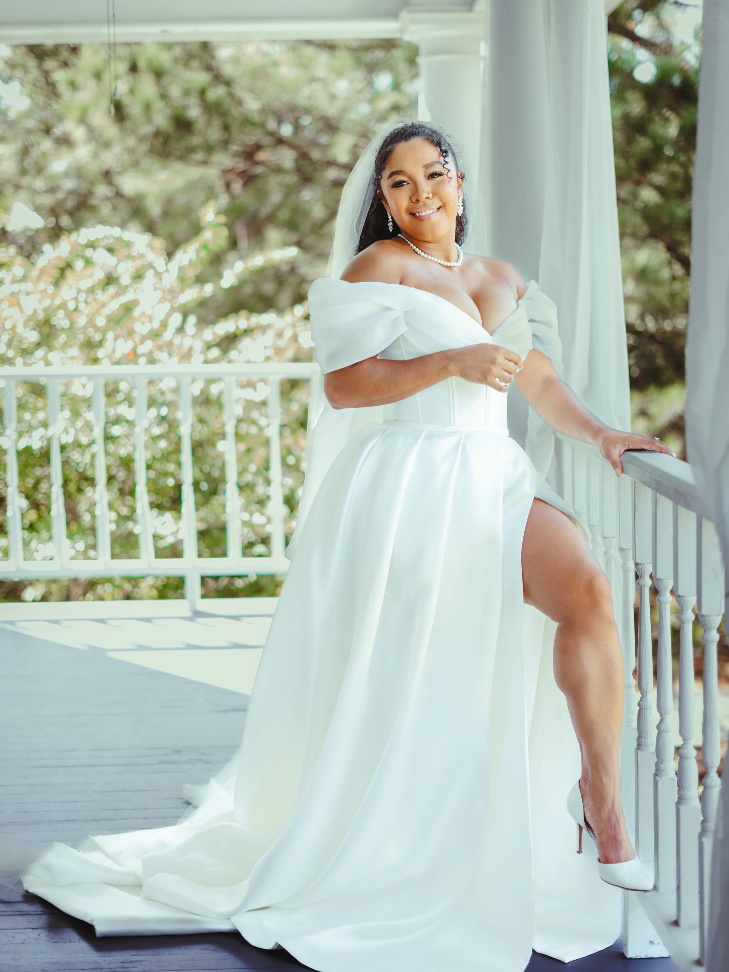 Bride in a white wedding dress standing on a porch, with off-the-shoulder sleeves and a pearl necklace and earrings, smiling at the camera.