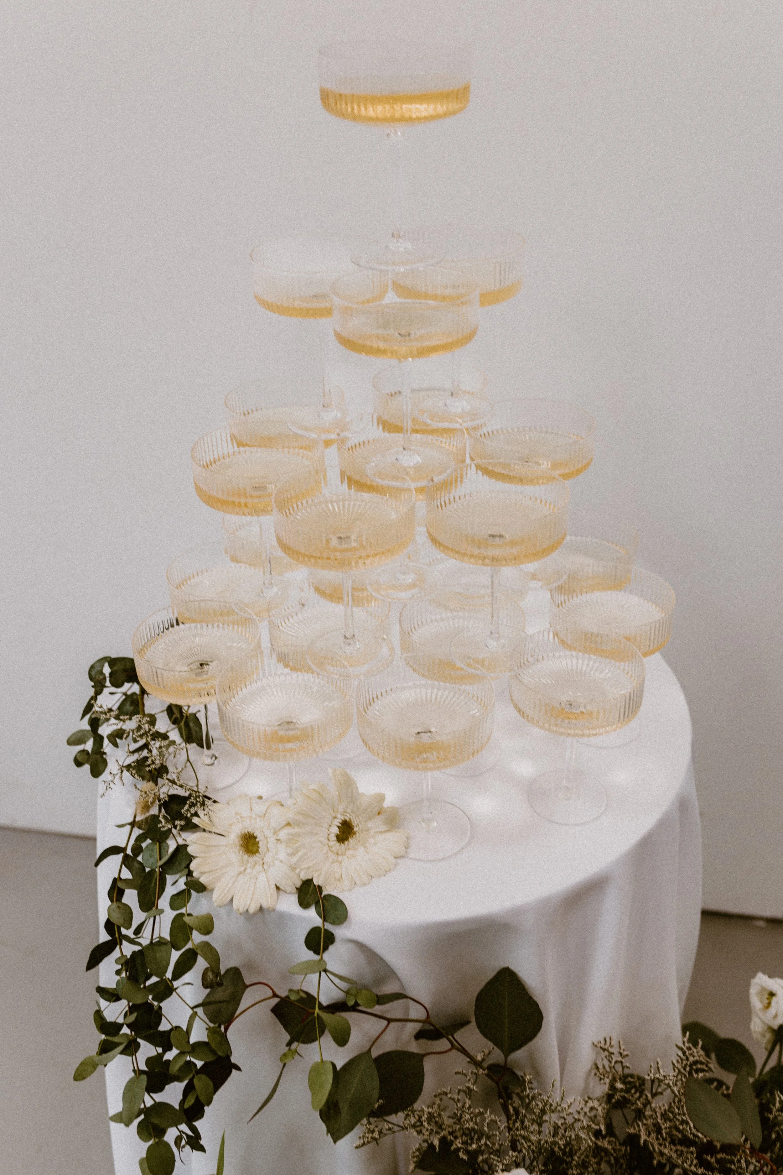 Champagne tower on a white tablecloth with green foliage and white flowers.