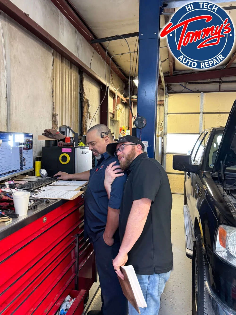 Two men smiling and talking inside an auto repair shop, with a red toolbox, computer, and automotive tools on the workbench, and a black vehicle parked nearby.