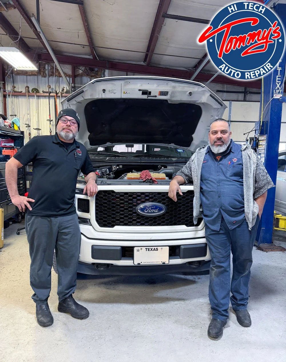Two mechanics standing in front of a white Ford vehicle with the hood open inside an auto repair shop.