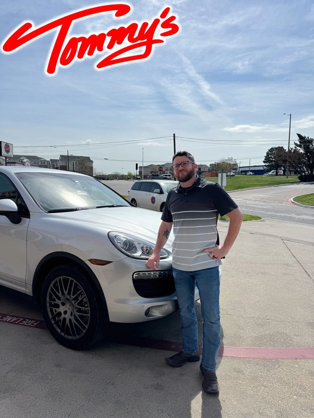 A man with glasses, a beard, and tattoos on his arms, standing next to a silver Porsche Macan.