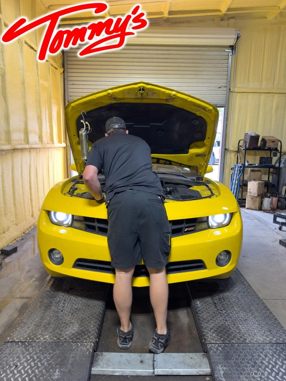 A person working on a yellow Chevrolet Camaro with the hood open inside an auto repair shop.