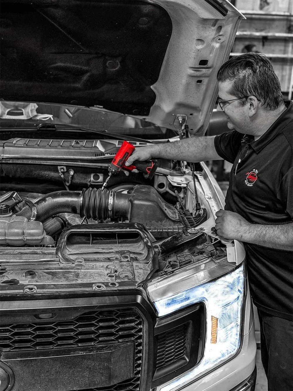 A mechanic working on a car engine in a garage, using a red cordless drill, with the hood open and the front of the vehicle visible.