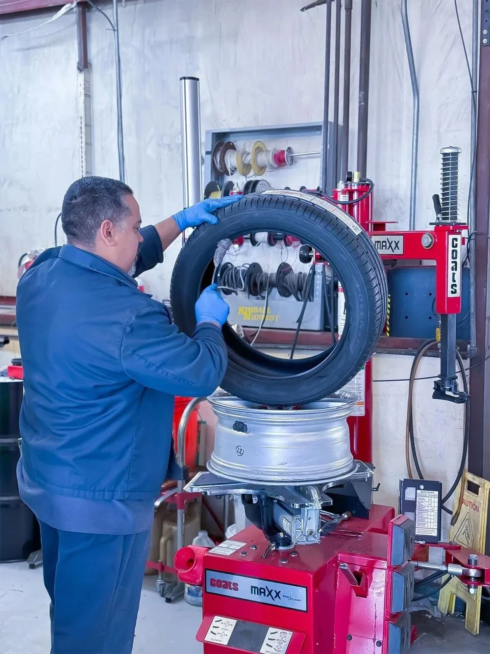 A mechanic wearing blue gloves and a blue jacket, working on a tire mounted on a red machine in a workshop, with various tools and spools of wire in the background.