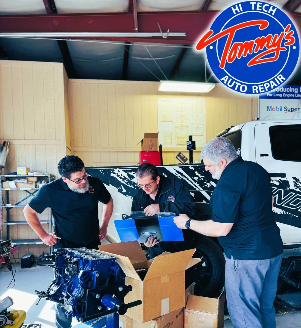 Three men work together in a garage, inspecting car parts and documents near a partially assembled blue engine, with a white and black vehicle in the background.