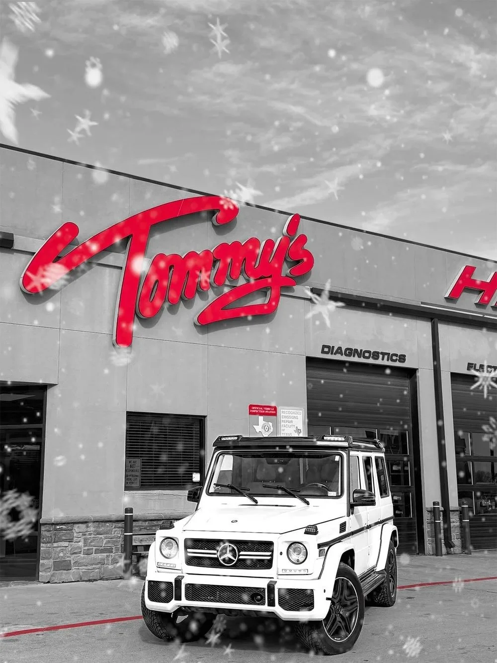A white Mercedes-Benz G-Class SUV parked outside a Tommy's Hi Tech Auto in Denton. The dealership's red illuminated sign is visible on the building. Snowflakes are falling, and the sky appears overcast.