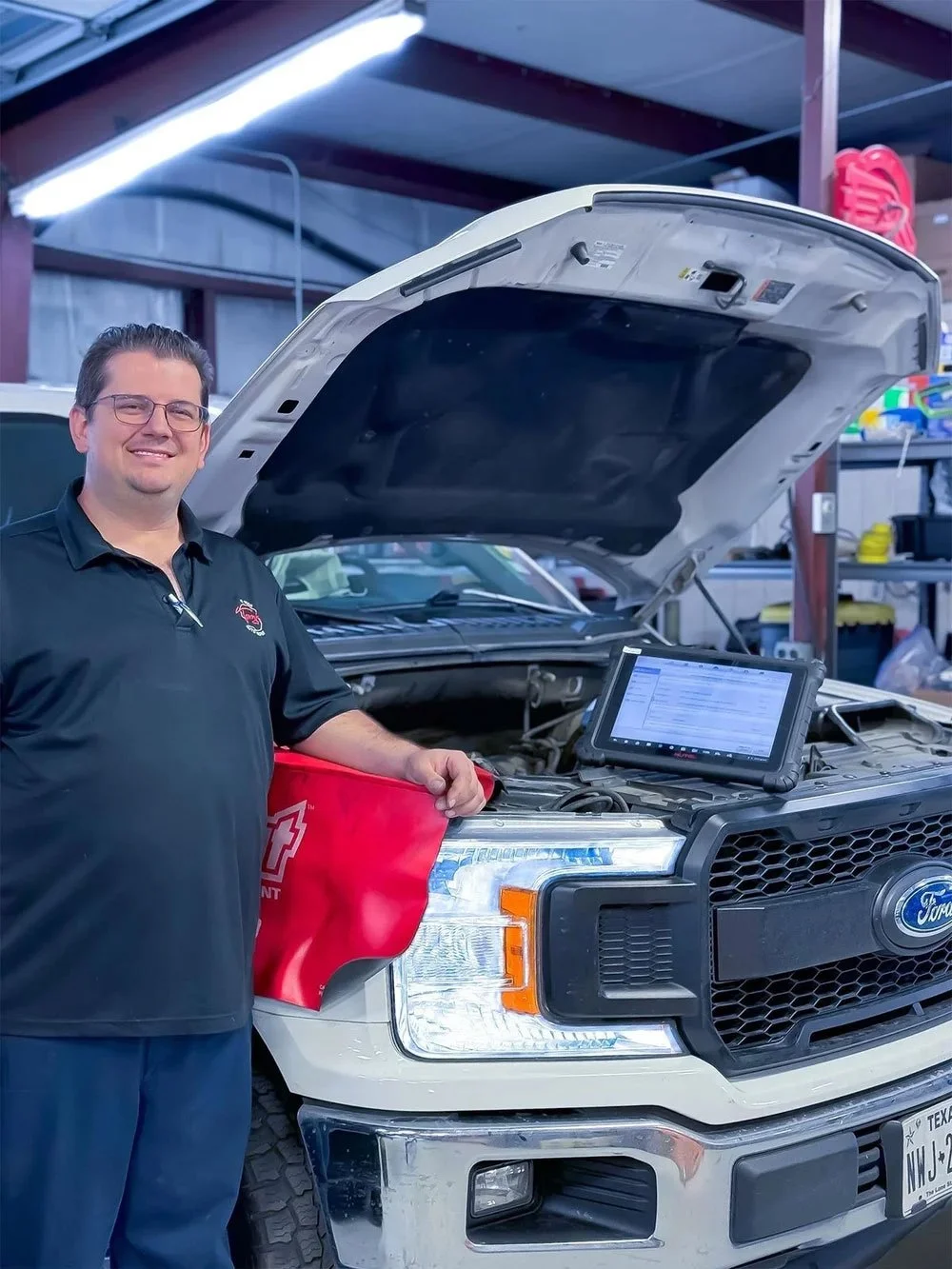 A mechanic standing next to a white Ford truck with the hood open, inside a garage. The mechanic is wearing a black polo shirt and glasses, smiling, and there is a tablet on the truck's engine showing diagnostic information.