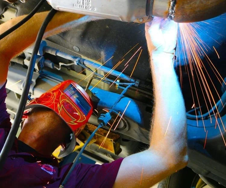 A worker welding metal components underneath a vehicle, wearing a red welding helmet and protective gloves, sparks flying from the welding process.