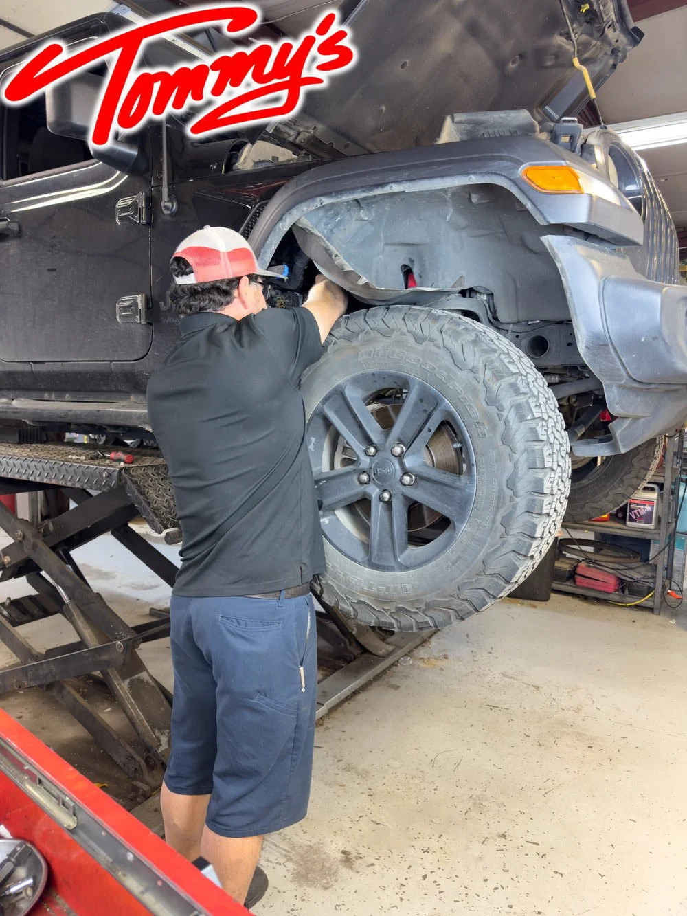 A mechanic works underneath a lifted black Jeep in a garage, with the vehicle's front wheel and fender visible, and the garage tools and shelves in the background.