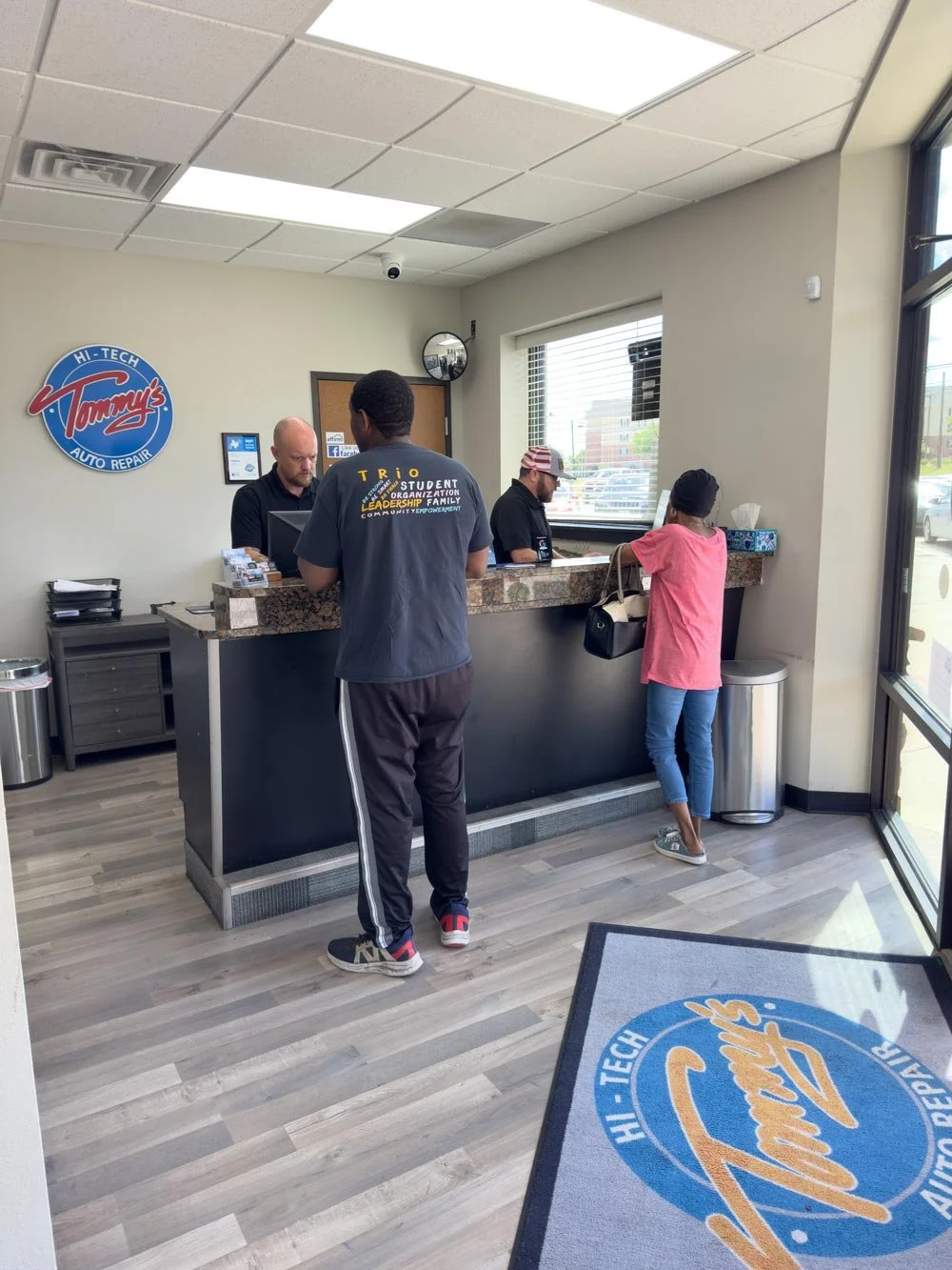 Customers standing at the counter inside Tommy's Hi-Tech Auto Repair shop, with employees assisting them. The shop has a wood-style floor, a large logo on the wall, and a branded mat on the floor near the entrance.