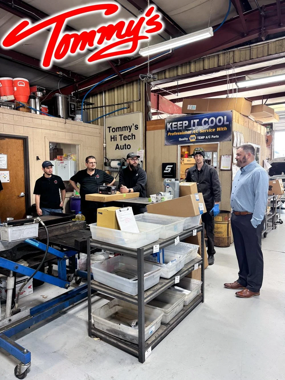 Group of five men having a discussion inside Tommys Hi Tech Auto garage, with equipment, boxes, and a sign reading 'Keep it Cool' in the background.