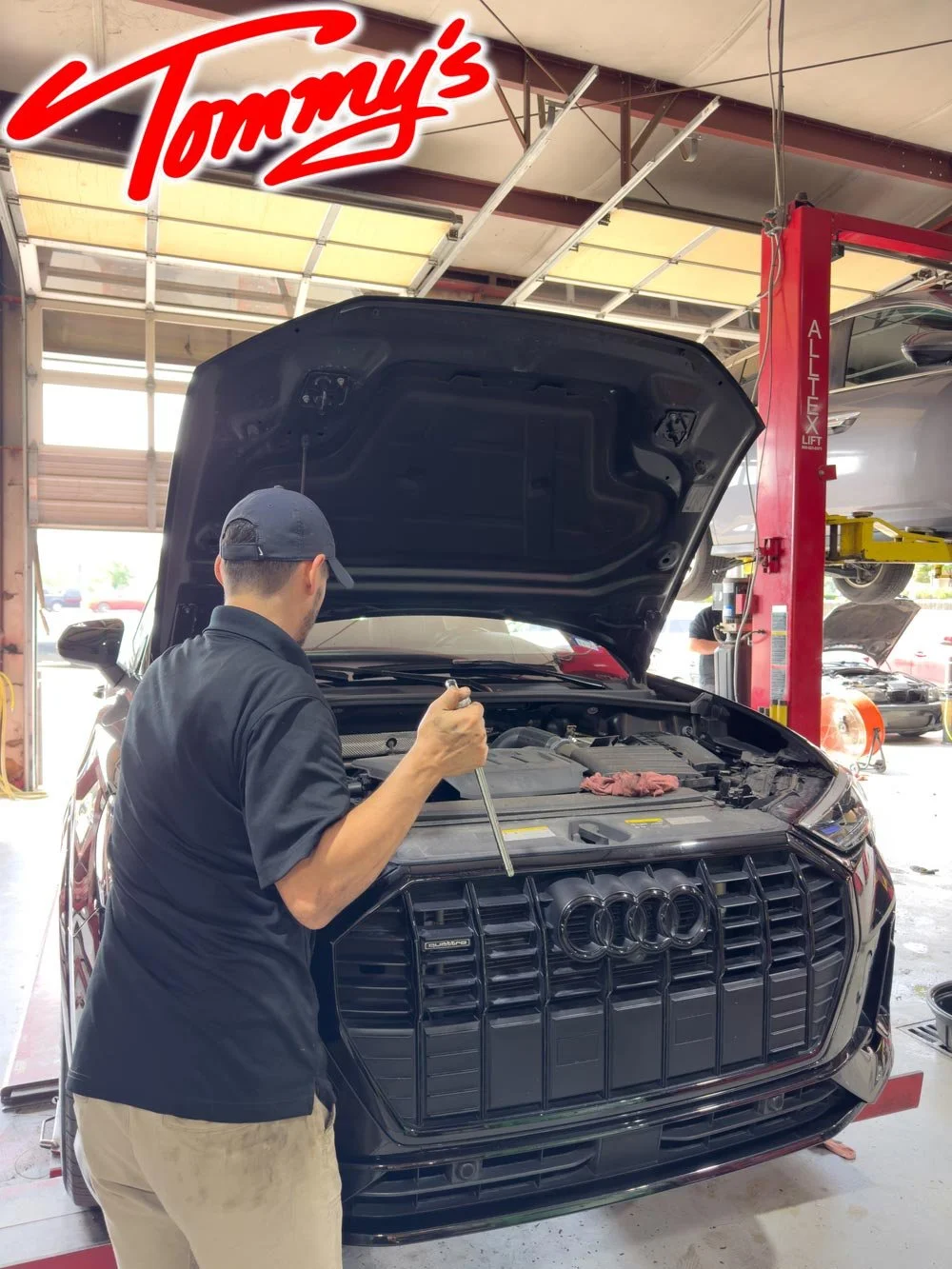 An auto mechanic working on the engine of a black Audi in a garage, with the garage door open and another car lifted in the background.