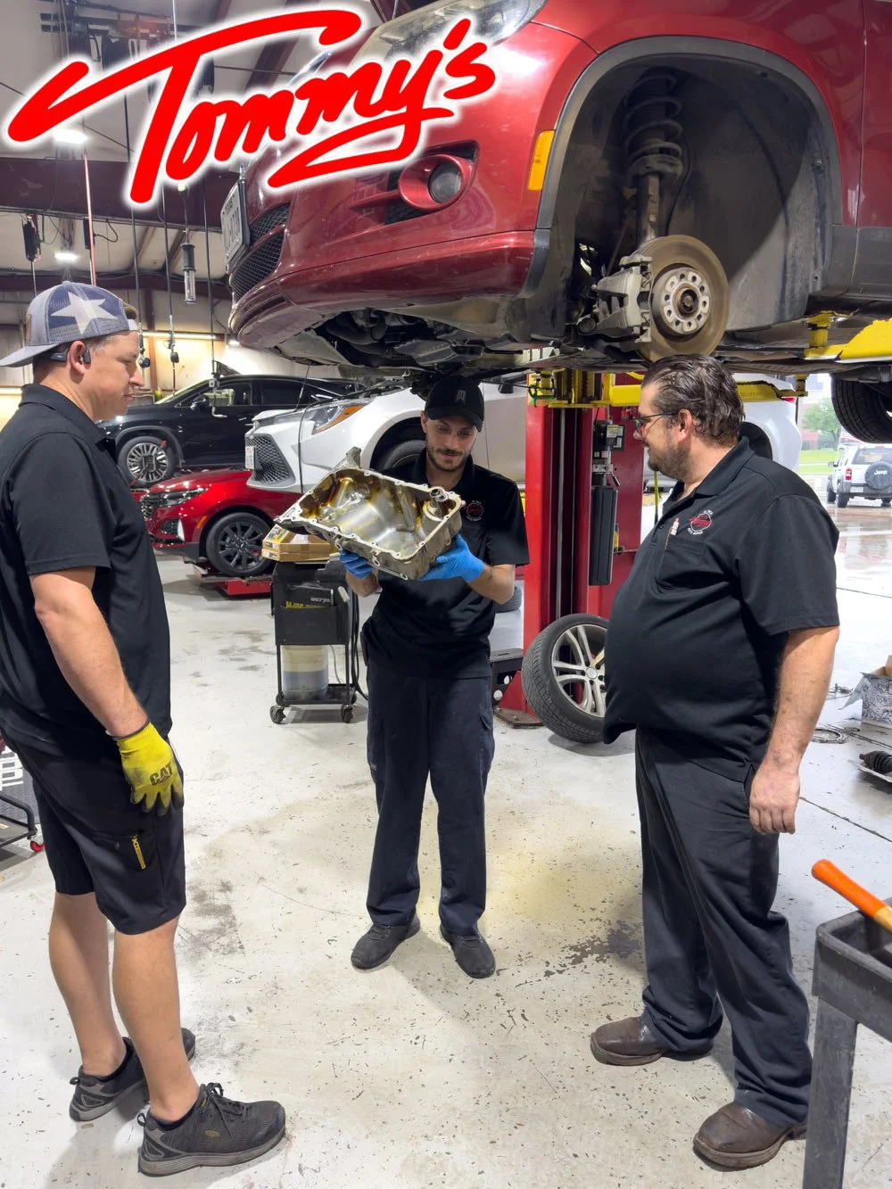 Three mechanics in black uniforms discussing engine parts in an auto repair shop with a car lifted above them. Various cars are visible in the background.