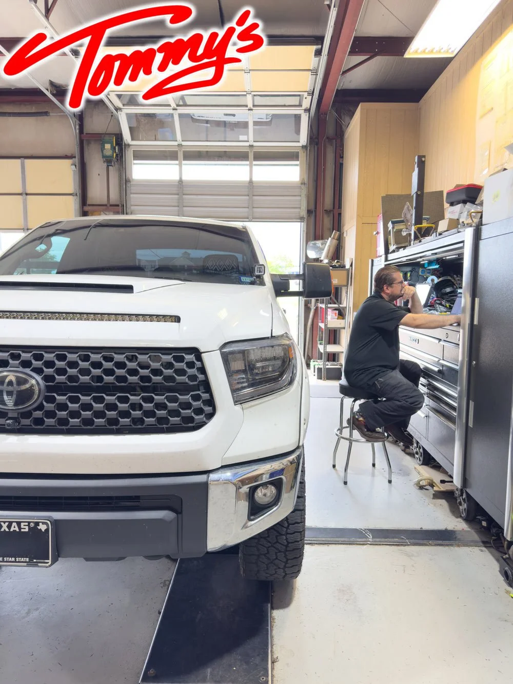 A white Toyota pickup truck is parked inside a garage, and a man in black work clothes is working at a large tool chest on the right side of the image.