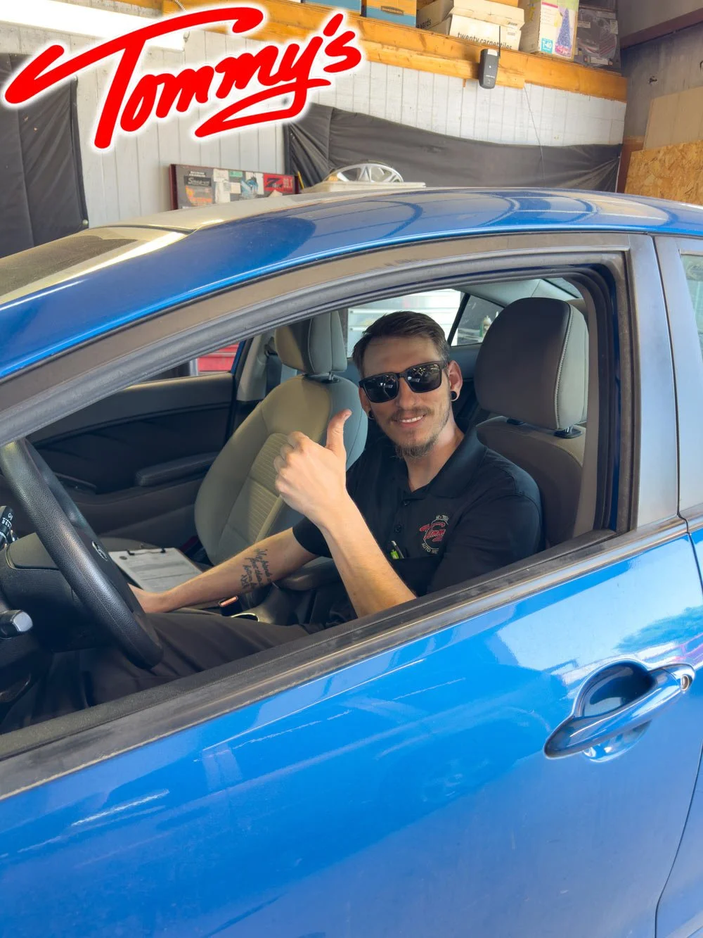 A man wearing sunglasses and a black polo shirt with a Tommy's logo is sitting in the driver's seat of a blue car, giving a thumbs-up and smiling inside a garage.