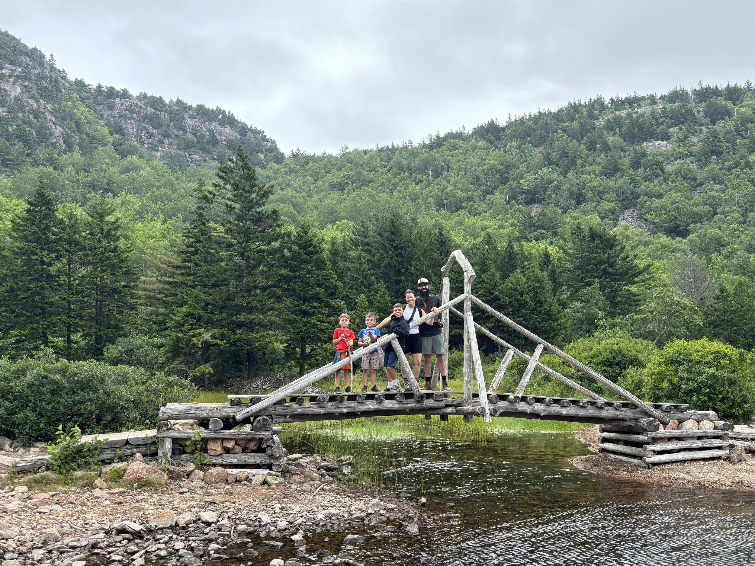 A group of six people, including children and adults, standing on a wooden bridge in a lush green forest with mountains in the background.