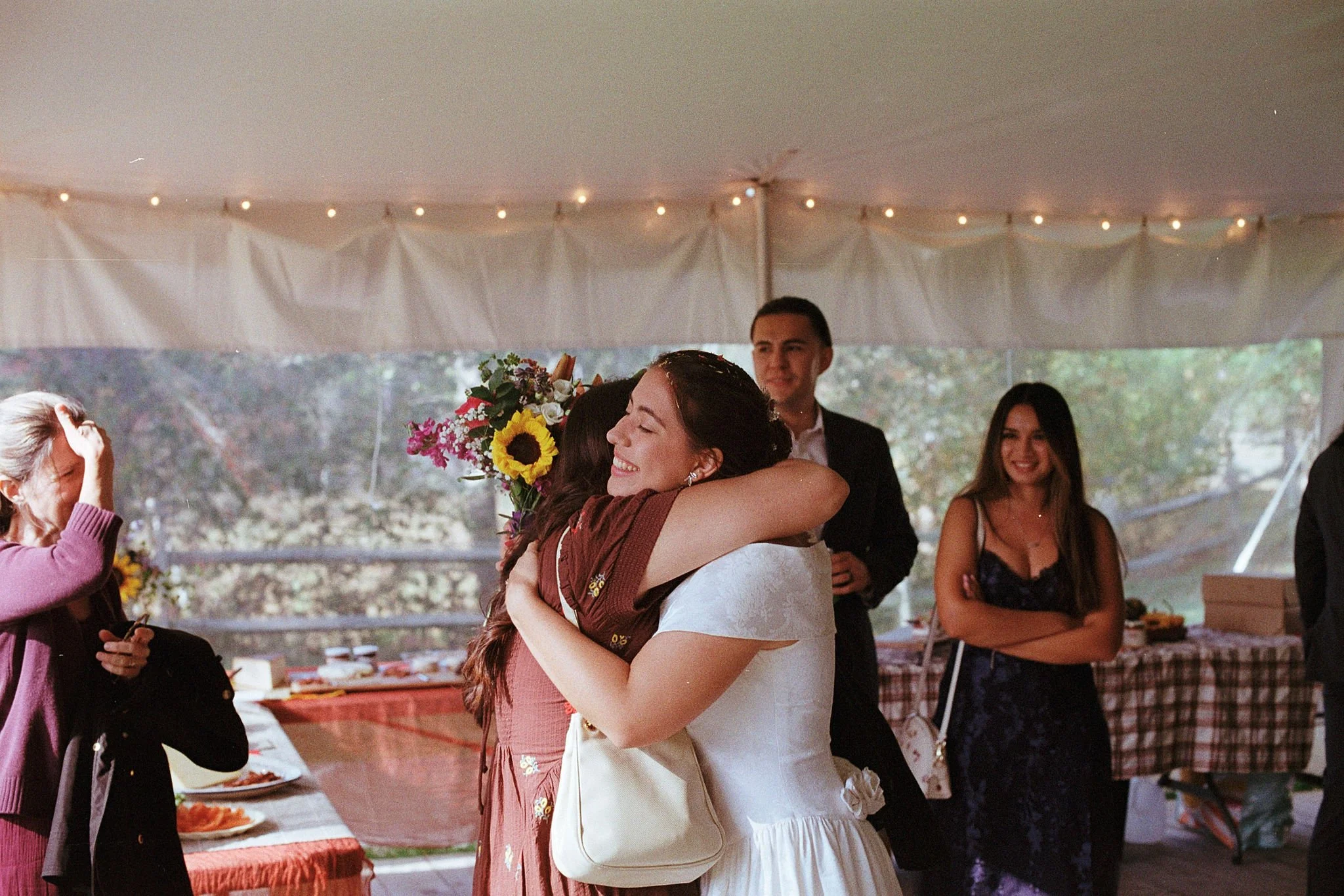 A bride holding a bouquet of flowers embraces her friend, who is wearing a brown dress. The bride is smiling with her eyes closed. A few guests stand around them, smiling. Taken at Red Ridge Farm in Massachusetts.