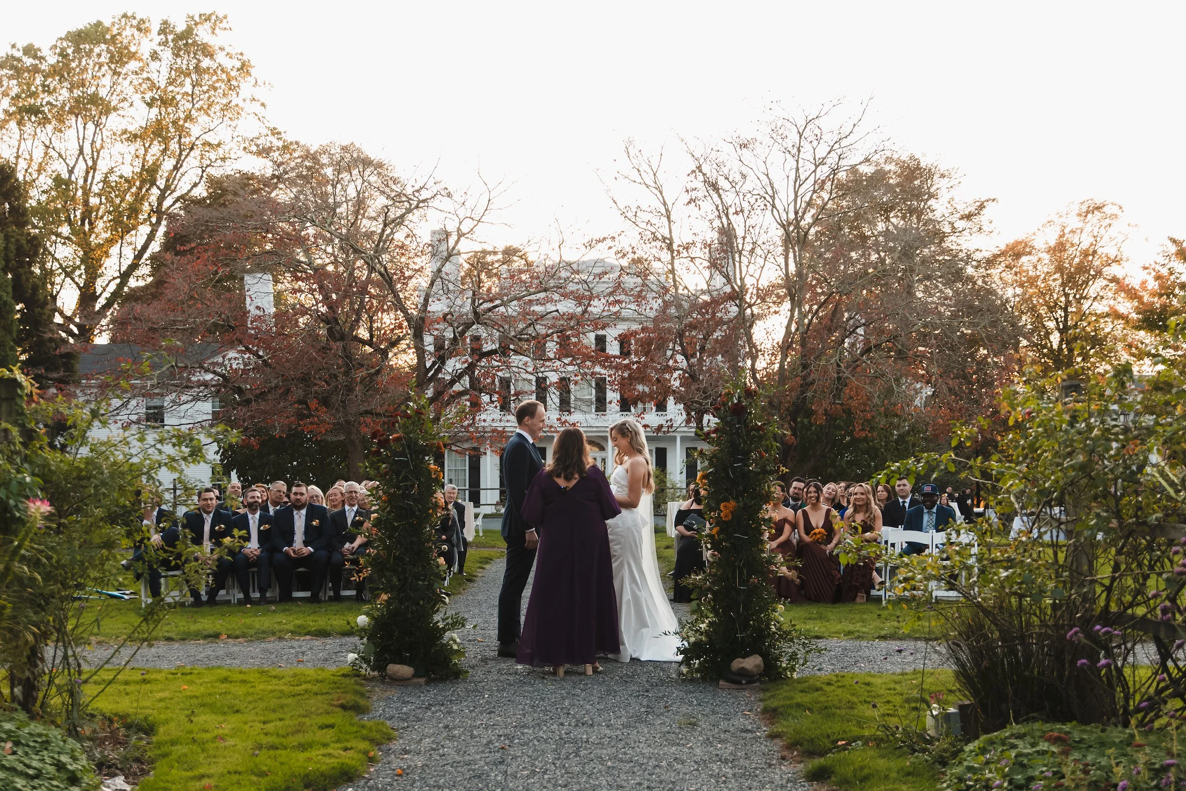 A bride and groom exchange vows amongst red foliage. A large white Colonial house looms in the background. Taken at Linden Place in Rhode Island.
