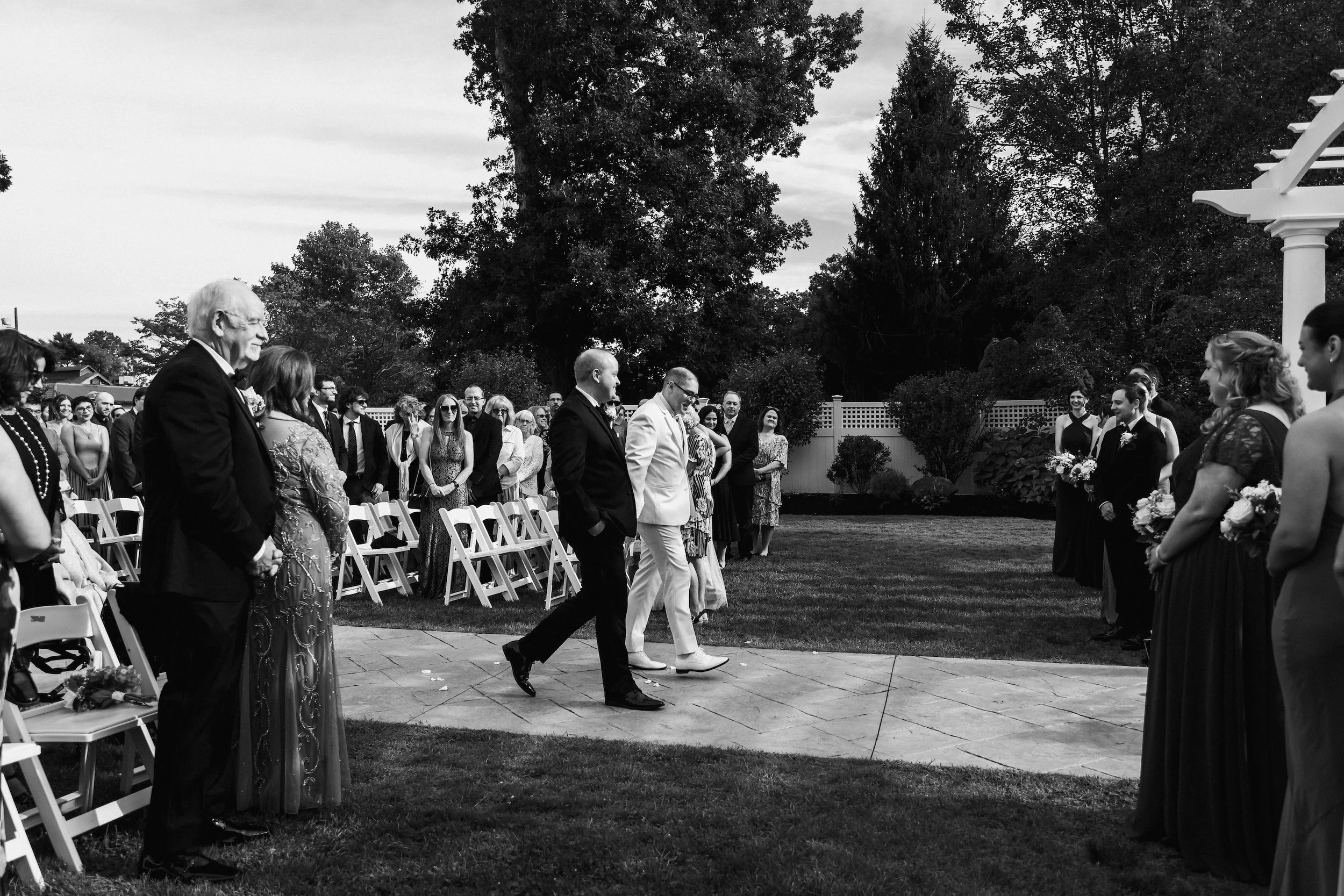 A profile view of two grooms walking toward a wedding arbor. One is dressed in a black suit, one is in a white suit. Their guests are seated and standing around them, smiling. Taken at The Villa At Ridder Country Club.