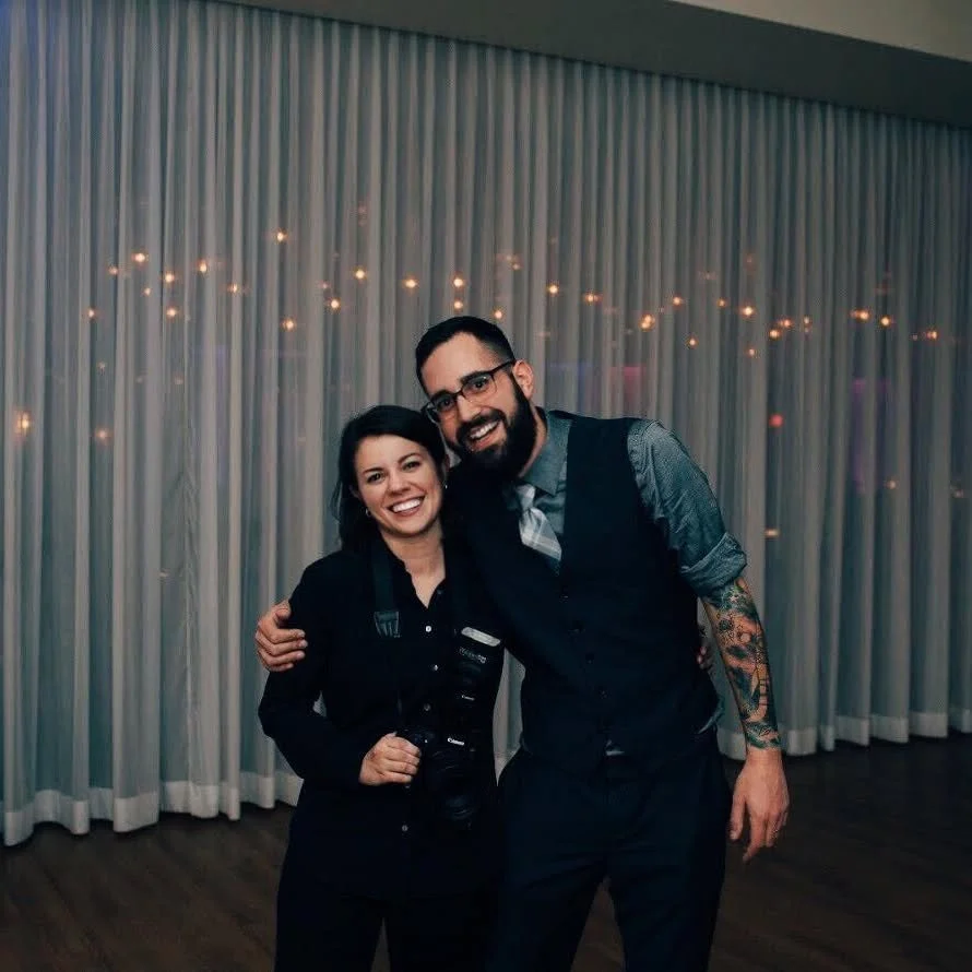Two people, a woman and a man, smiling and posing arm-in-arm indoors with a curtain and string lights in the background.