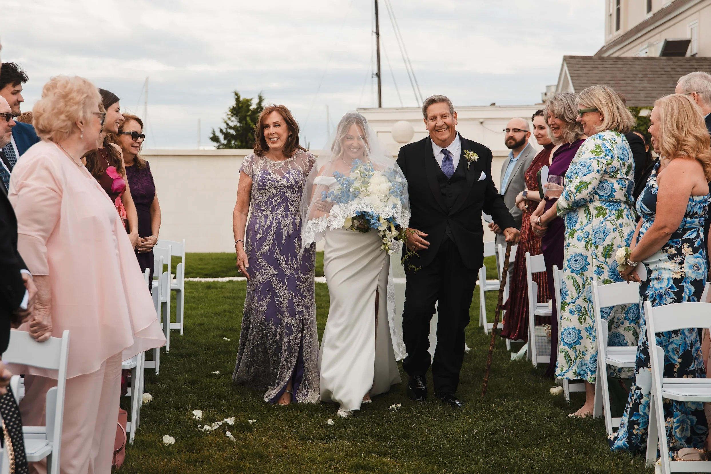 A veiled bride is walking down an aisle toward the arbor with her parents on her side. All are smiling. Guests surround them on the aisle. Taken at Belle Mer in Newport, Rhode Island.
