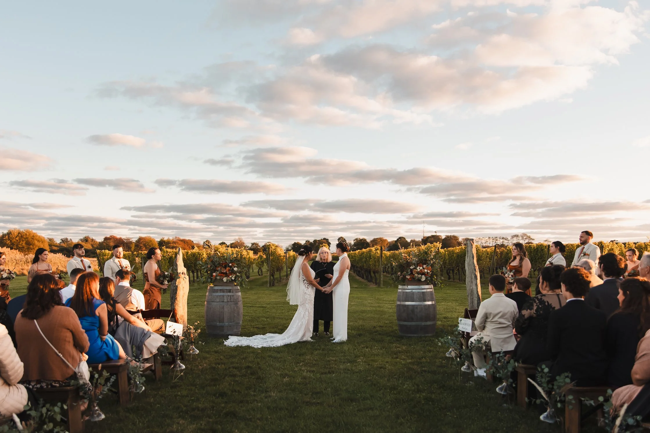 Two brides exchange vows in front of rows of grapevines at sunset. Family and friends are seated in rows in front of them. Taken at Newport Vineyards in Newport, Rhode Island.