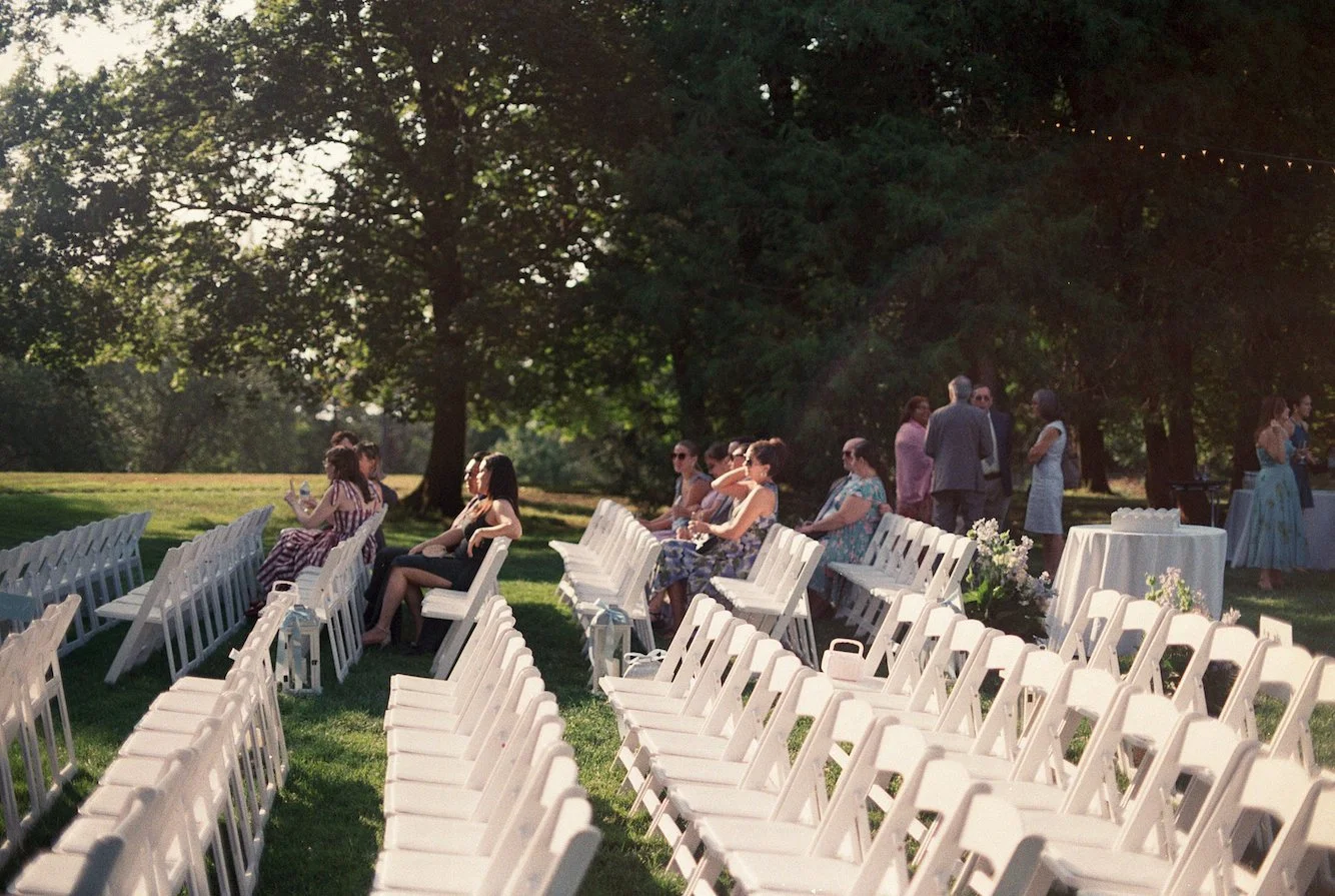 Outdoor wedding reception setup on a grassy lawn with rows of white chairs, some people seated and others standing, under large trees with string lights. Taken at Stevens Estate.
