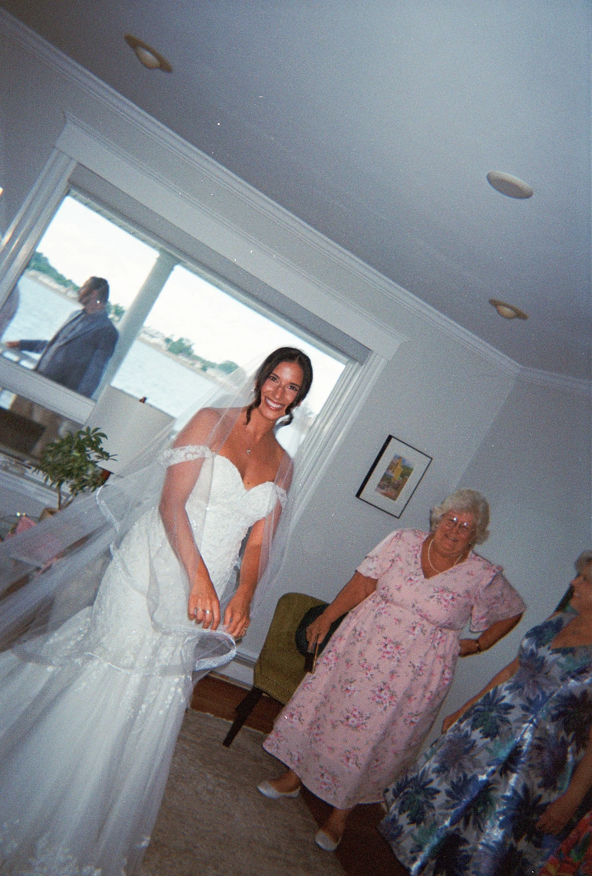 A bride wearing a white dress and veil smiles at the camera inside a home. A woman in a pink dress stands behind her. Taken on private property in Hull, Massachusetts
