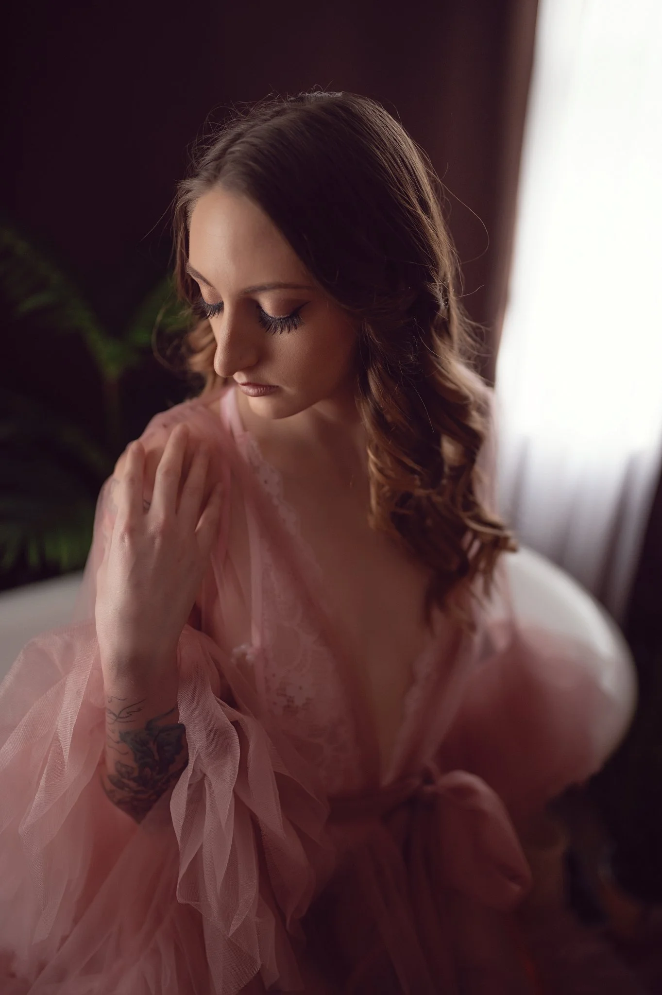 A woman with long, wavy brown hair and makeup, wearing pink lace lingerie and a sheer pink robe, sitting by a window with light coming through, some greenery in the background.