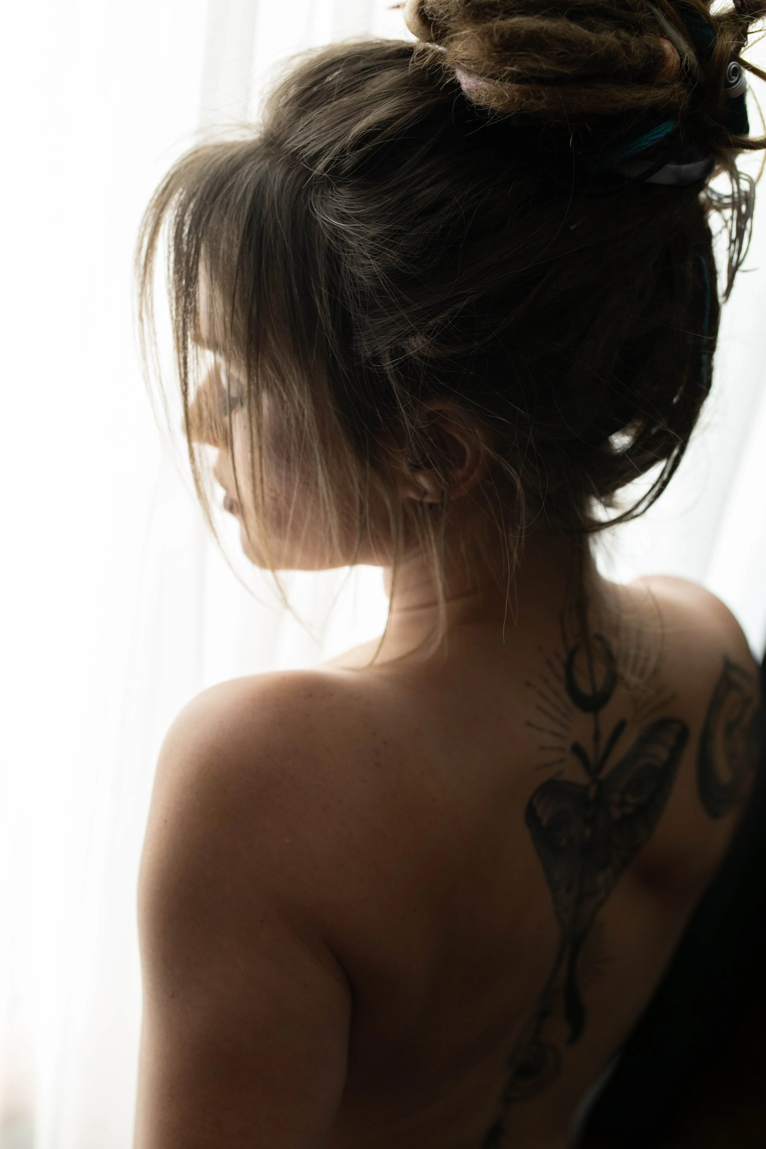 A woman with tattoos on her shoulder and back, with shadowed hair styled in dreadlocks, sitting in front of a bright, white background.