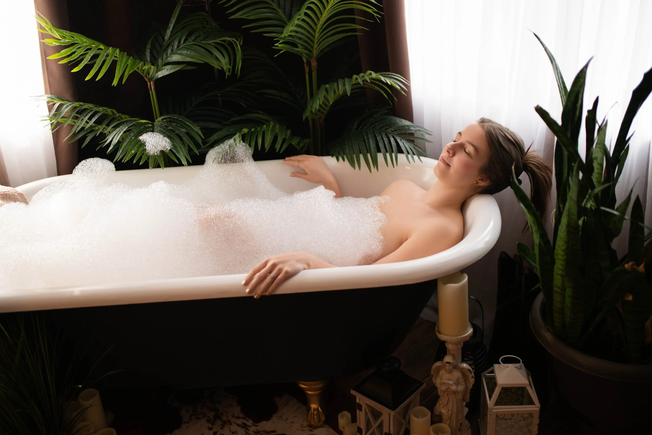 A young woman relaxing in a bathtub filled with bubbles, surrounded by green plants and candle holders.