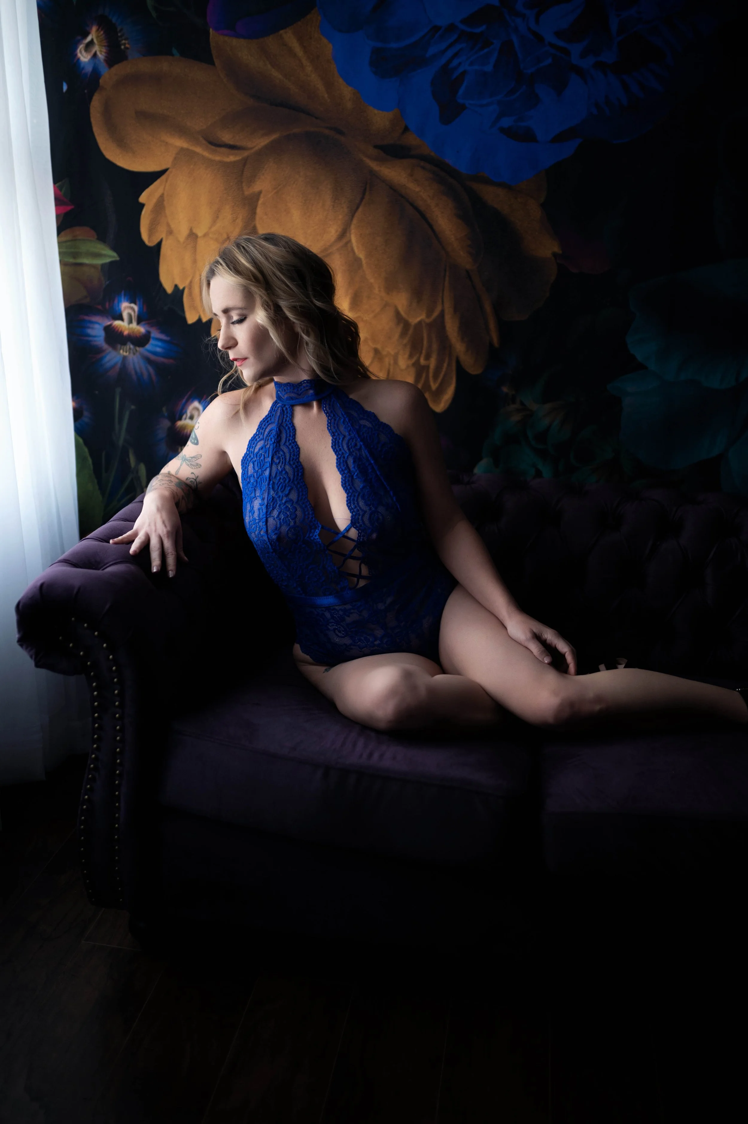 A woman in a blue lace lingerie sitting on a dark purple velvet sofa, with a floral wall mural in the background and natural light coming from the left.