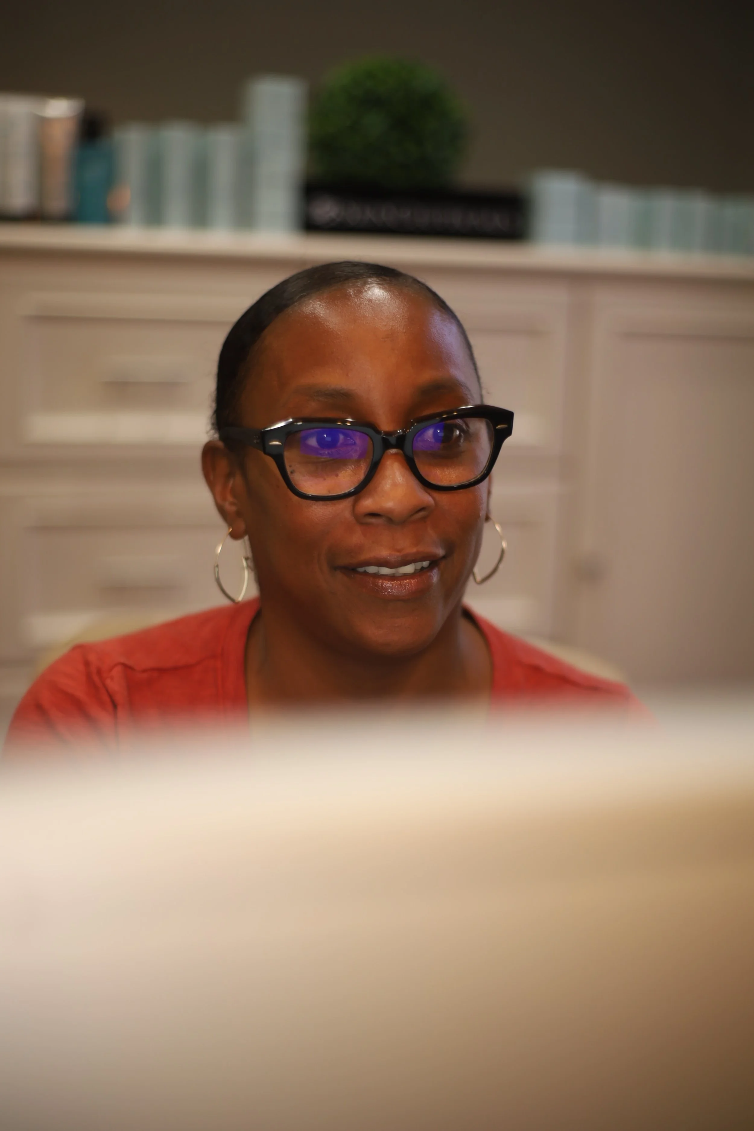 A woman with short dark hair, glasses with purple reflections, and hoop earrings, smiling while working at a desk in an indoor setting.