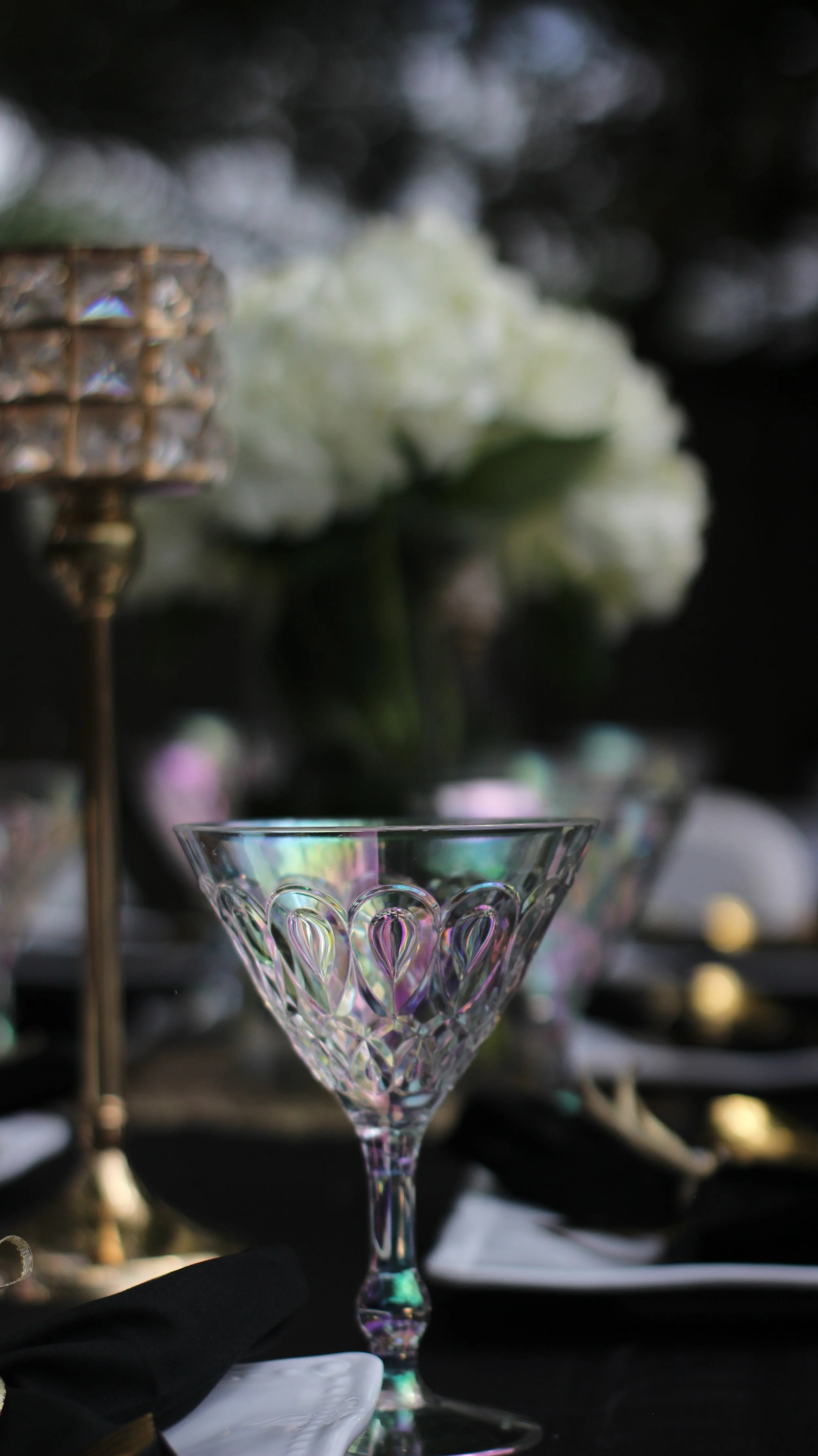 An elegant glass dessert coupe with intricate heart-shaped cutouts, placed on a table with black napkin, blurred white flowers in the background, and a decorative lamp on the left.