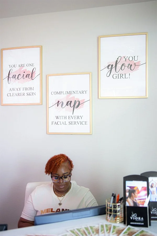 A woman with short, curly orange hair and glasses sitting behind a reception desk in a skincare or beauty salon. Behind her are three framed signs on a light-colored wall that promote facial services and skin health.