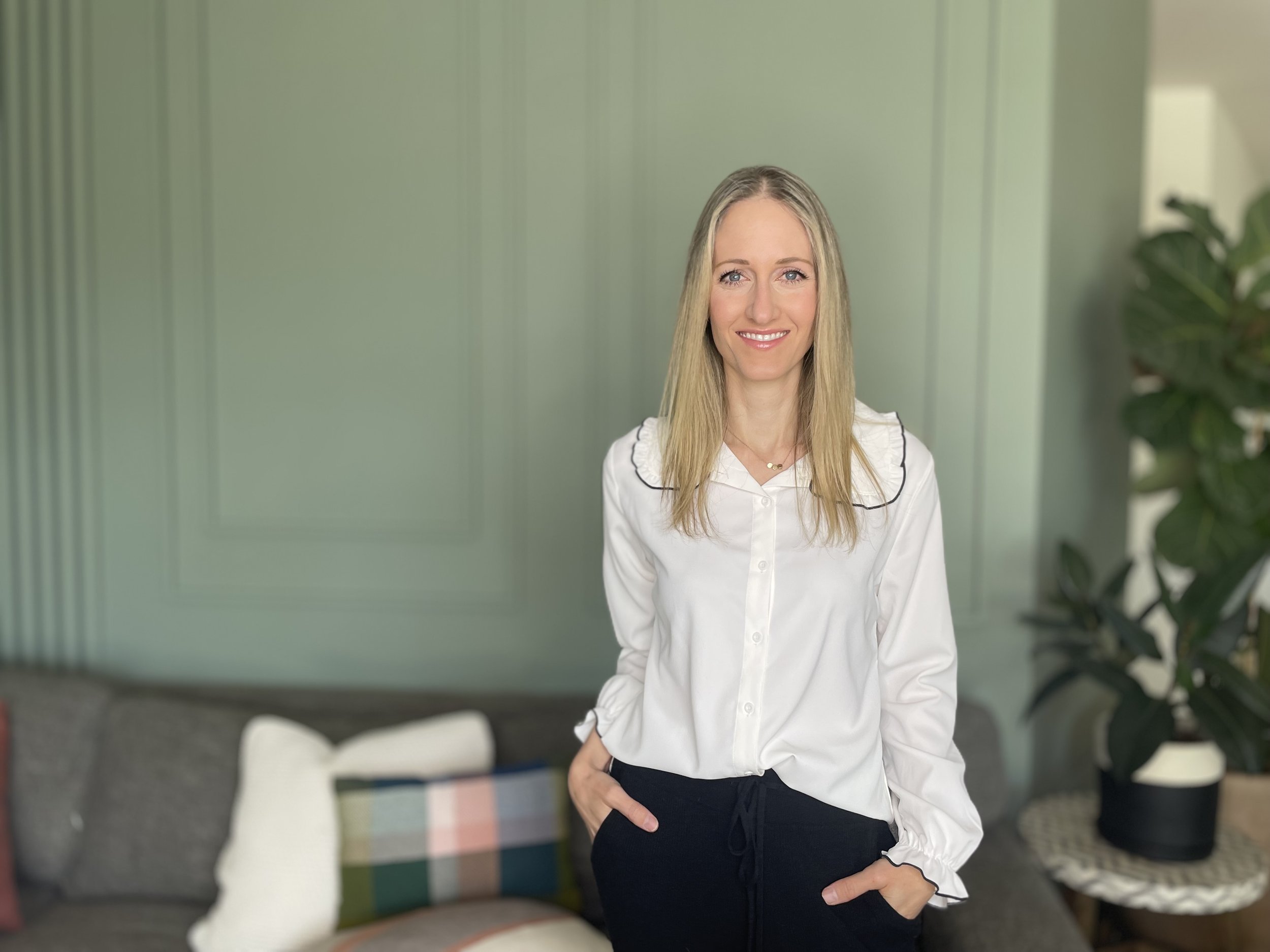 Marissa, owner of Fix My Space Marissa, wearing a white blouse and black pants, standing in a living room with a green wall and a large plant in the background.