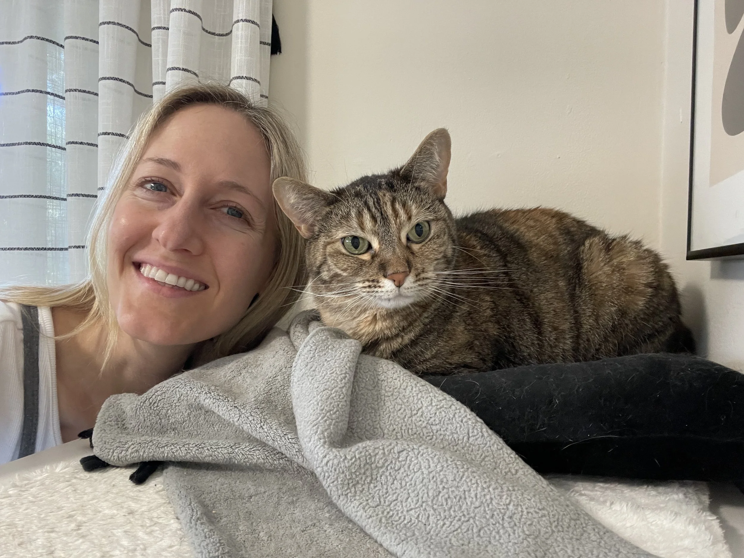 A woman with blonde hair smiling and lying on her side next to a tabby cat lying on a blanket, with a window and curtains in the background.