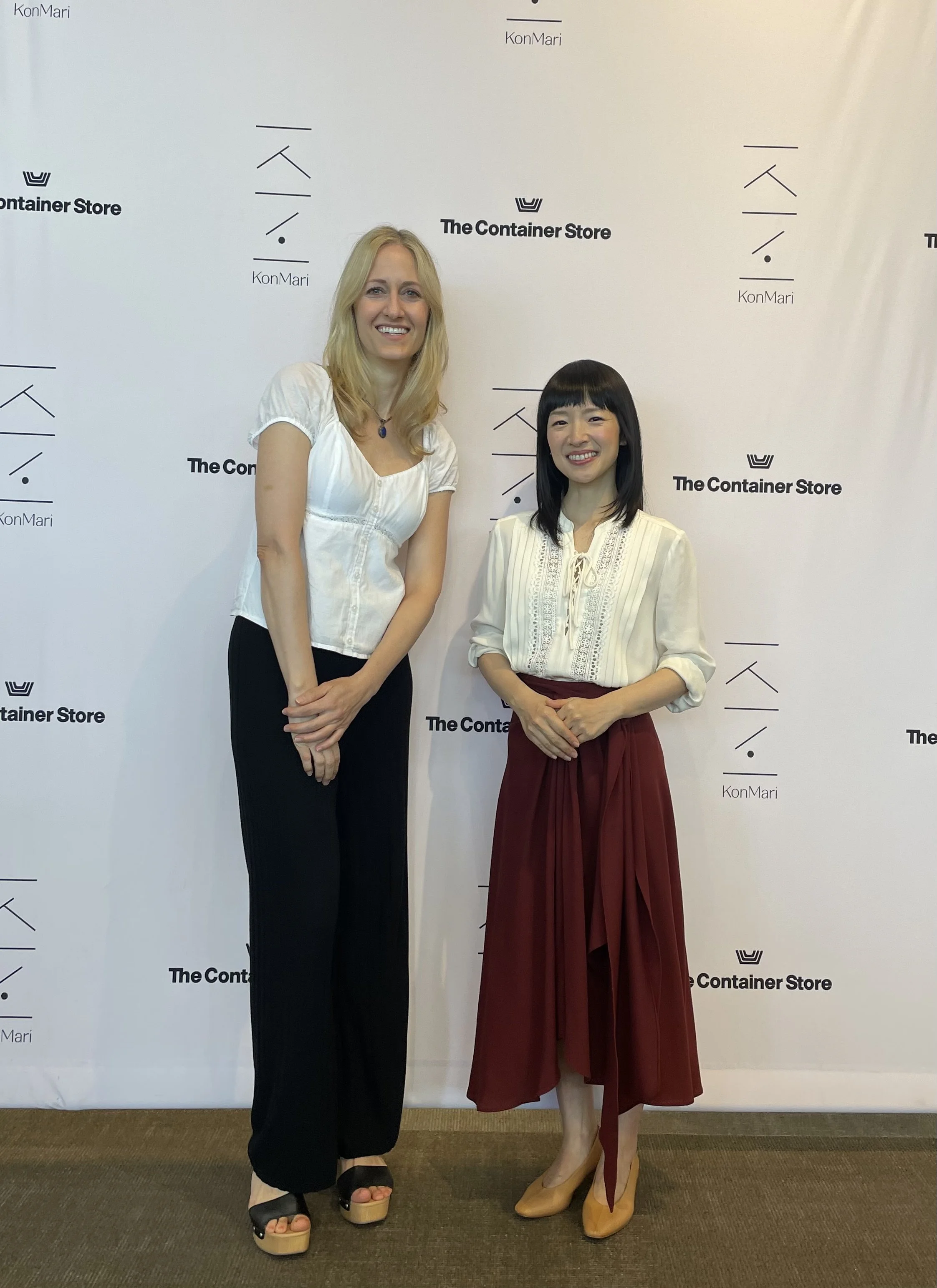 Two women standing in front of a white backdrop with logos from The Container Store and KonMari. The woman on the left is tall with blonde hair, wearing a white blouse, black pants, and black sandals. The woman on the right has black hair with bangs, wearing a cream blouse, a red skirt, and beige heels. Both are smiling.