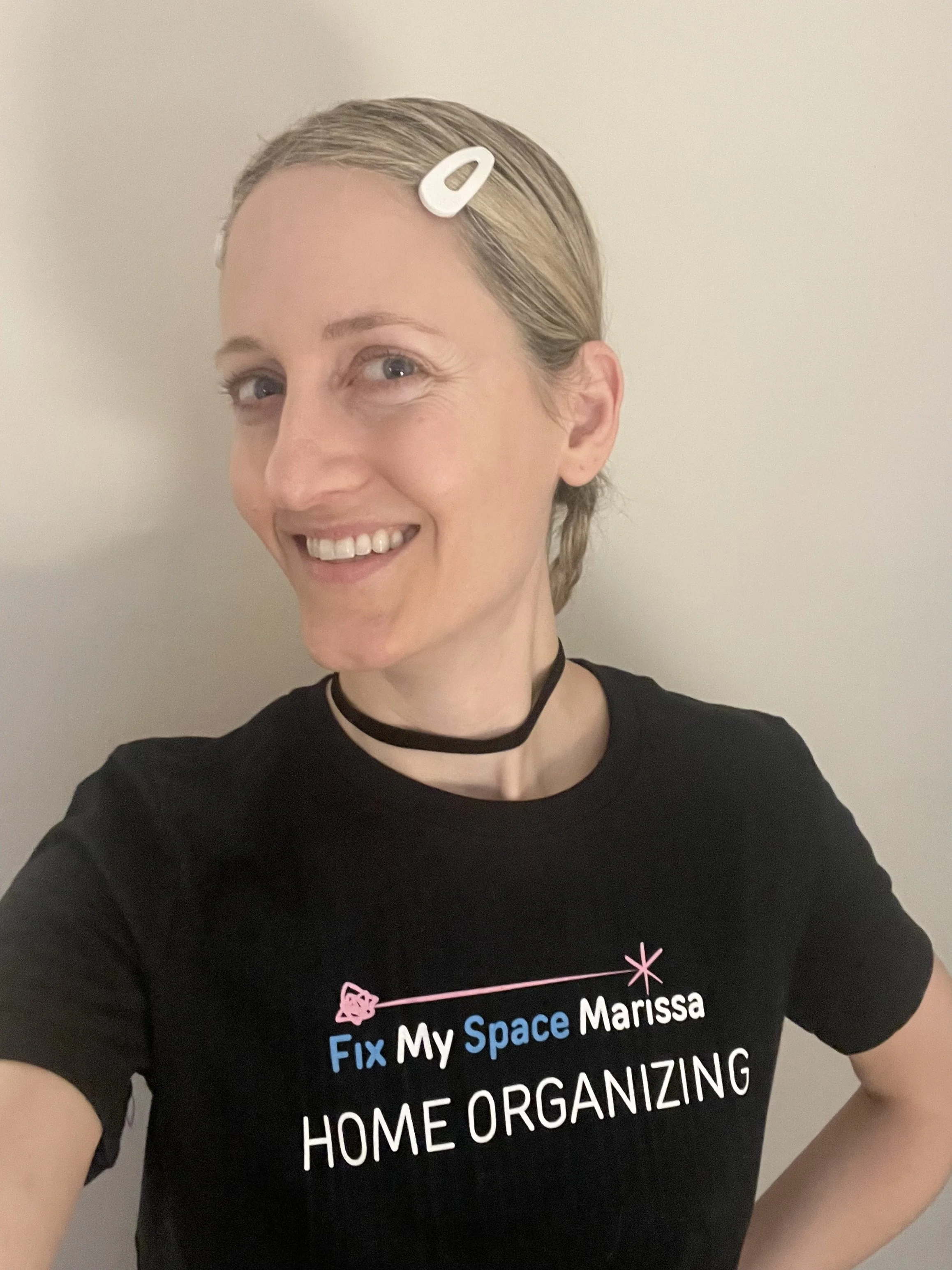A woman smiling and taking a selfie against a plain light-colored wall. She has blonde hair clipped back and is wearing a black T-shirt with text that says 'Fix My Space Marissa Home Organizing' and a black choker necklace.