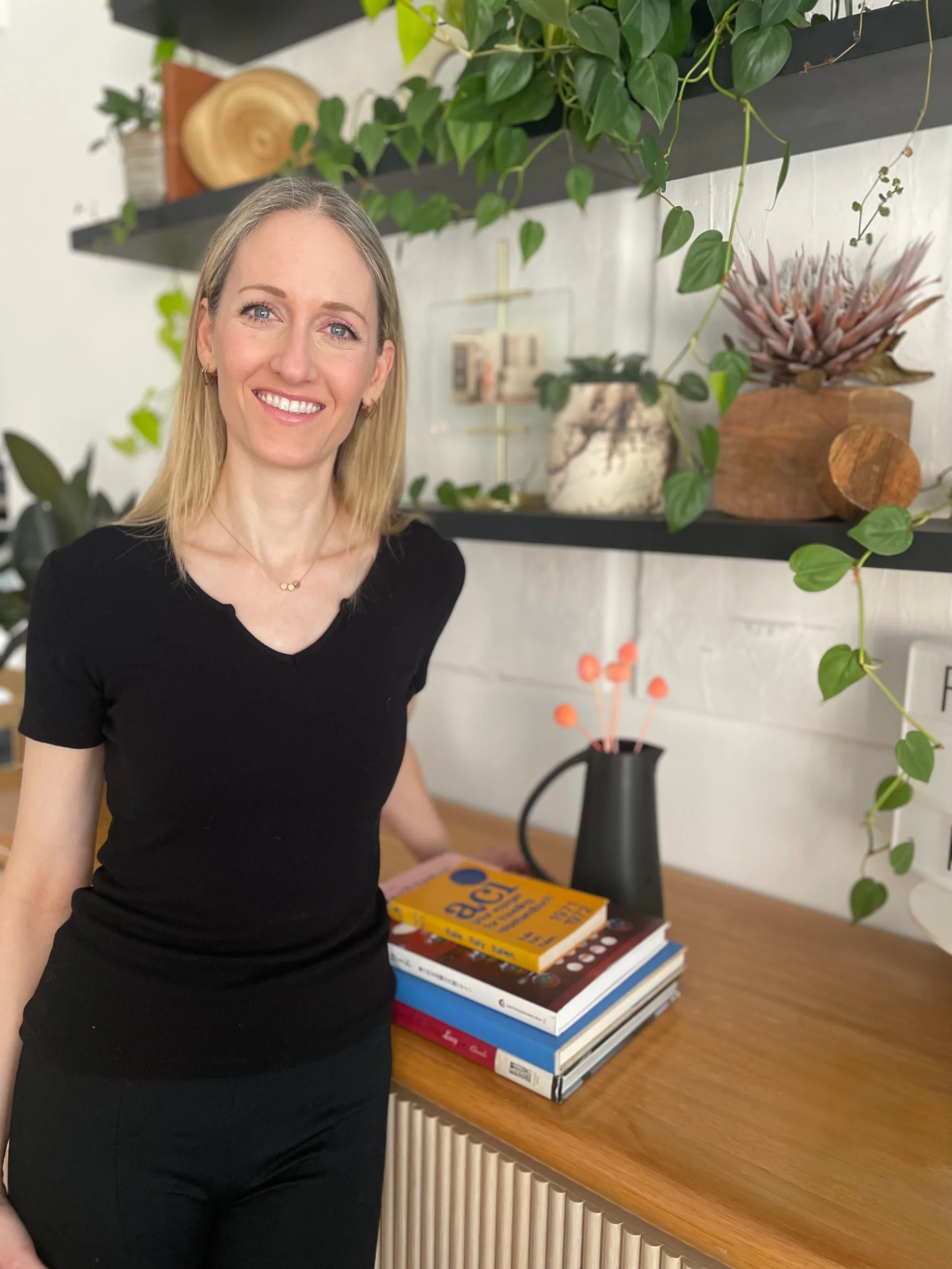 A woman with blonde hair wearing a black shirt and black pants stands in front of a wooden table with a stack of colorfully decorated books and a black vase with orange flowers. Behind her is a white brick wall with black shelves holding plants, a wooden bowl, and decorative items.