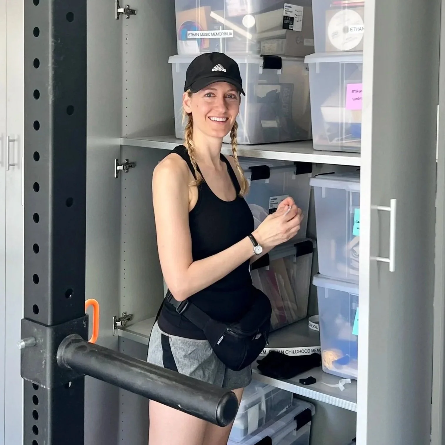 A young woman with blonde hair in two braids, wearing a black baseball cap, black tank top, and gray shorts, stands inside a storage cabinet with plastic bins, smiling at the camera.