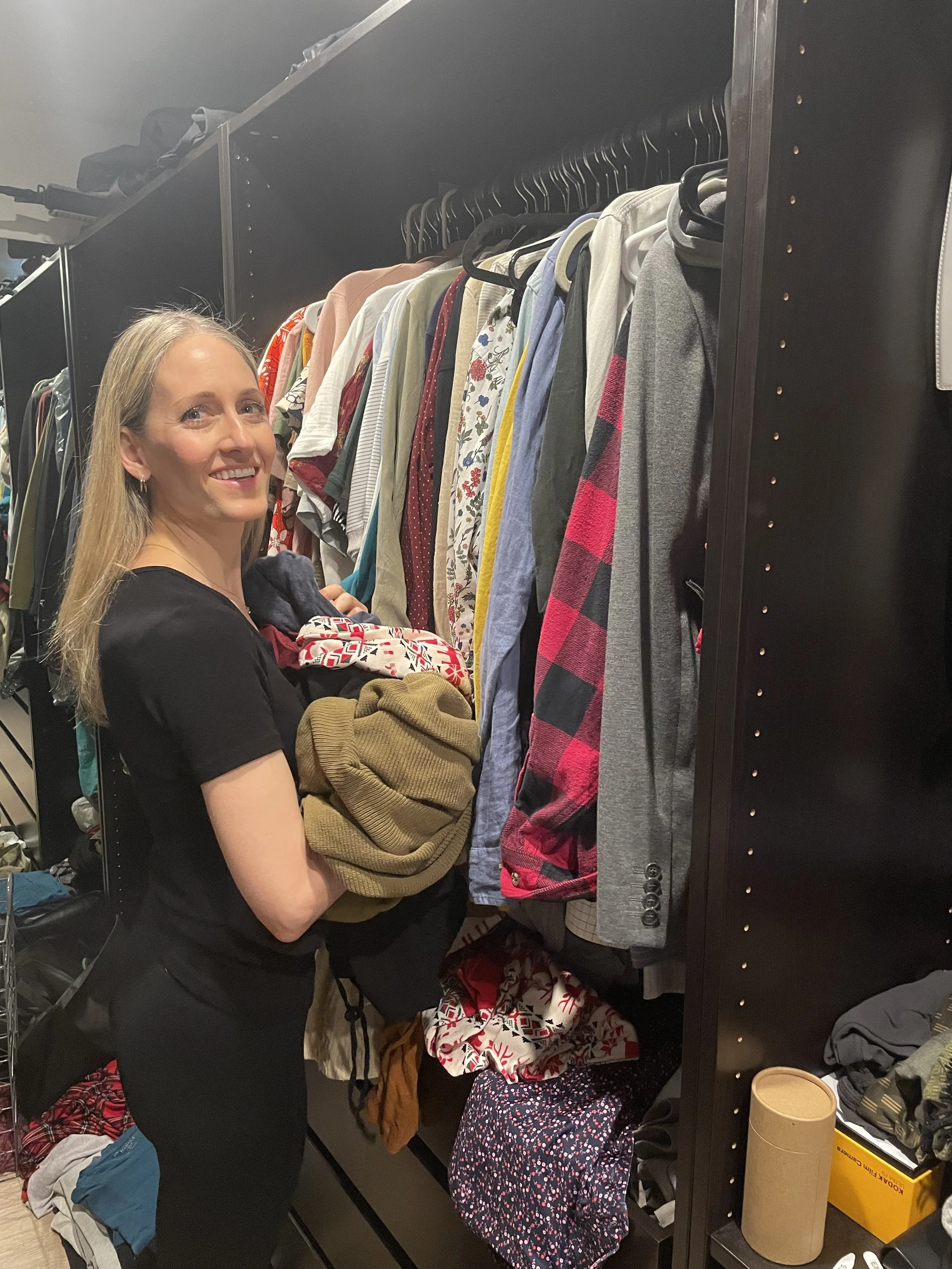 Marissa, owner of Fix My Space Marissa, shopping for clothes in a closet or dressing room, holding folded sweaters and surrounded by various hanging and folded clothes.