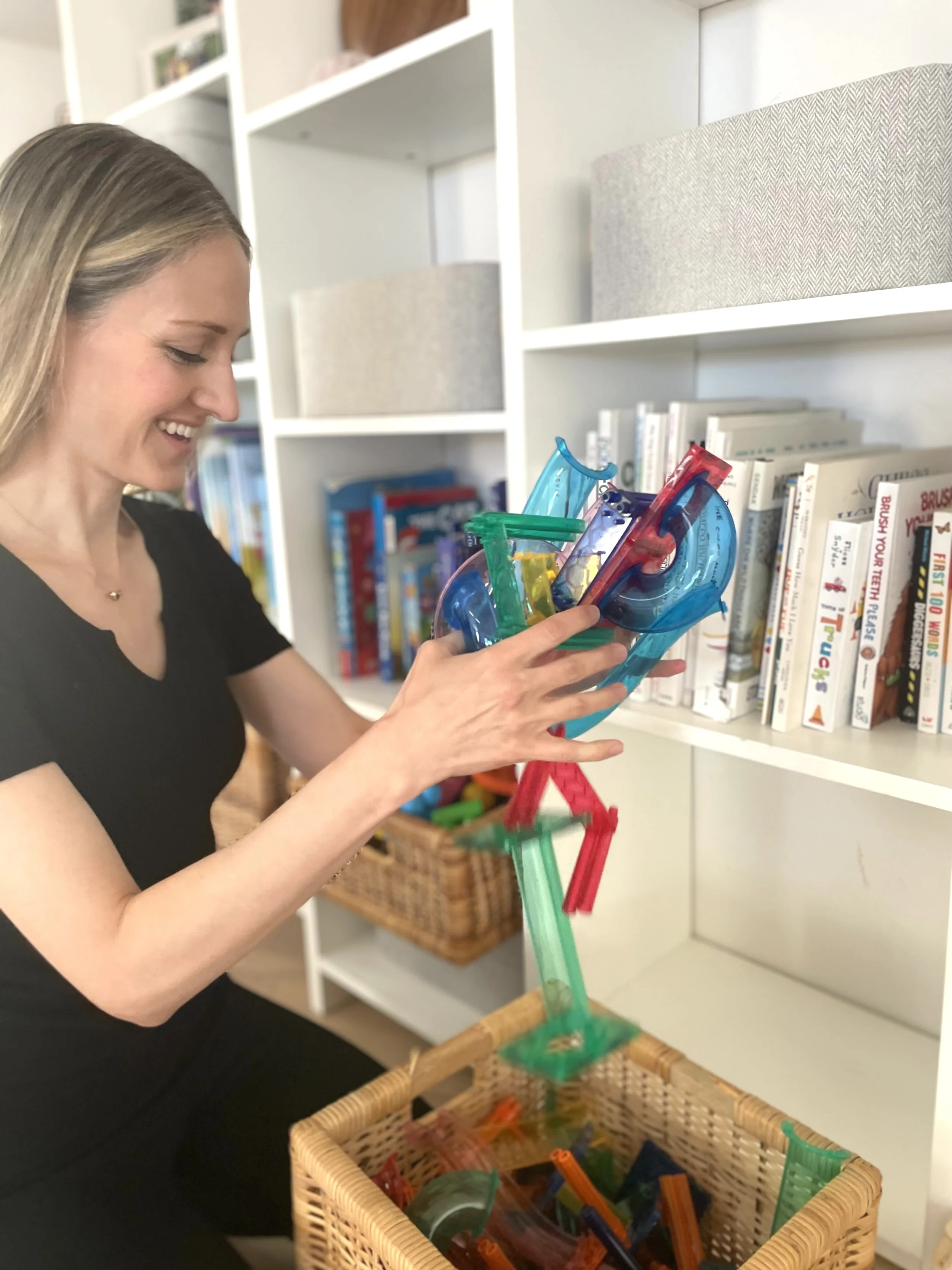 Marissa, owner of Fix My Space Marissa is playing with colorful straws, stacking and balancing them above a basket of more straws, in a room with bookshelves and storage bins.